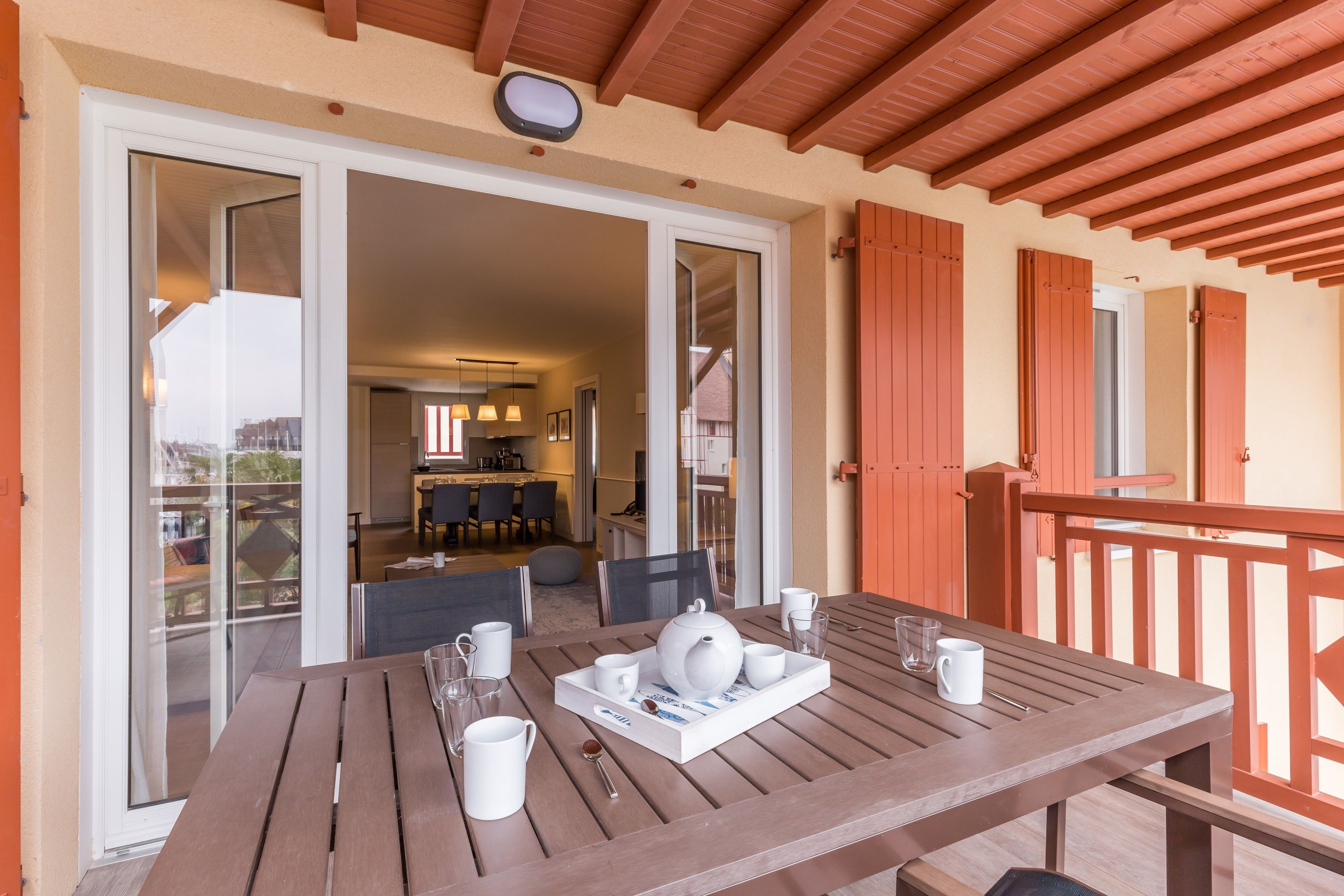 A wooden table set with white teapot, cups, and saucers on a balcony with orange shutters and a view of the interior.