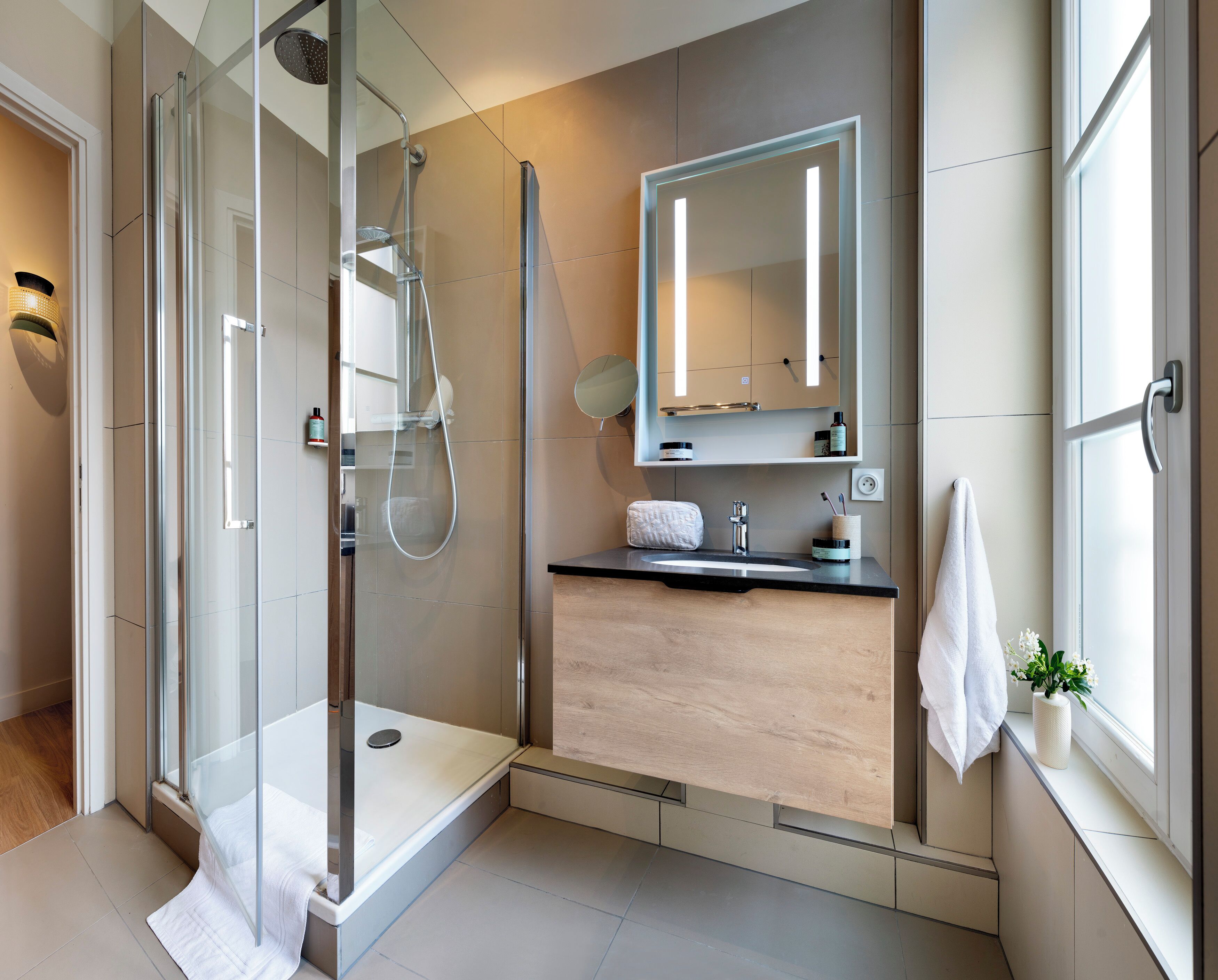 Een moderne badkamer met een douchecabine van glas, een houten wastafelmeubel, een grote spiegel en een raam voor natuurlijk licht.