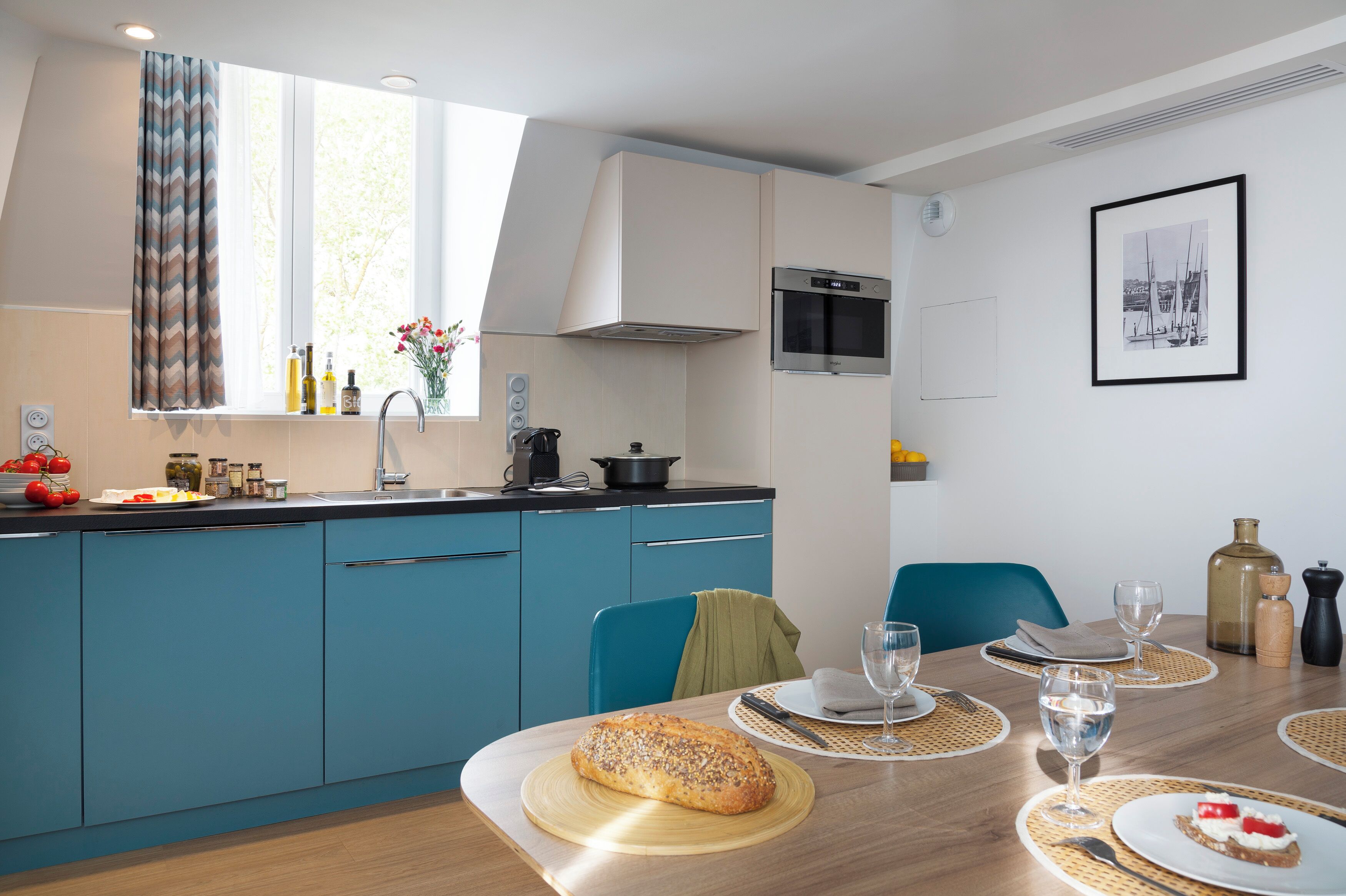 A modern kitchen with blue cabinets, a dining table set with plates, glasses, and a loaf of bread.