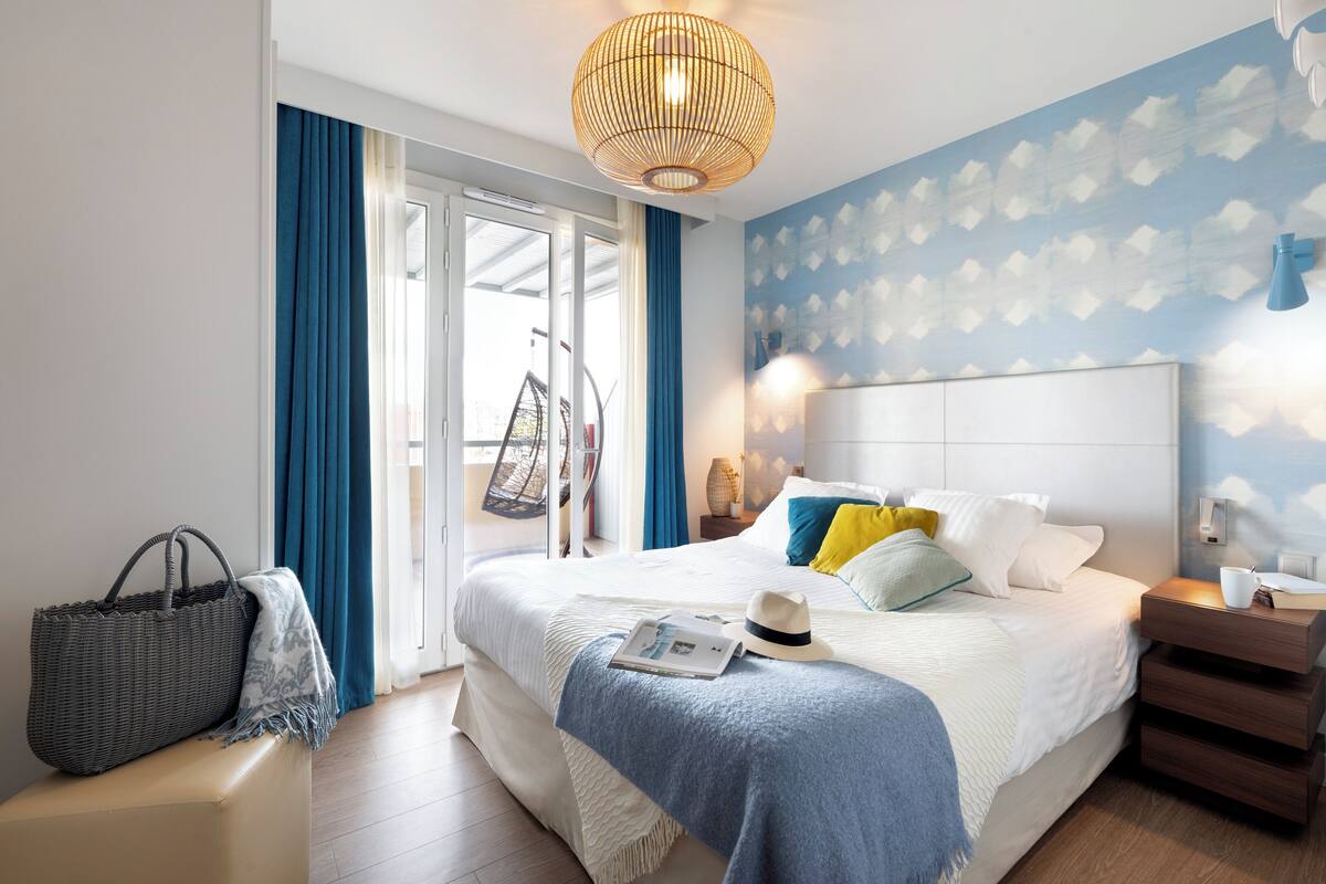 Quarto de hotel moderno com uma cama grande, ventoinha no teto, parede com padrão de nuvens e vista para o exterior através de uma porta deslizante.