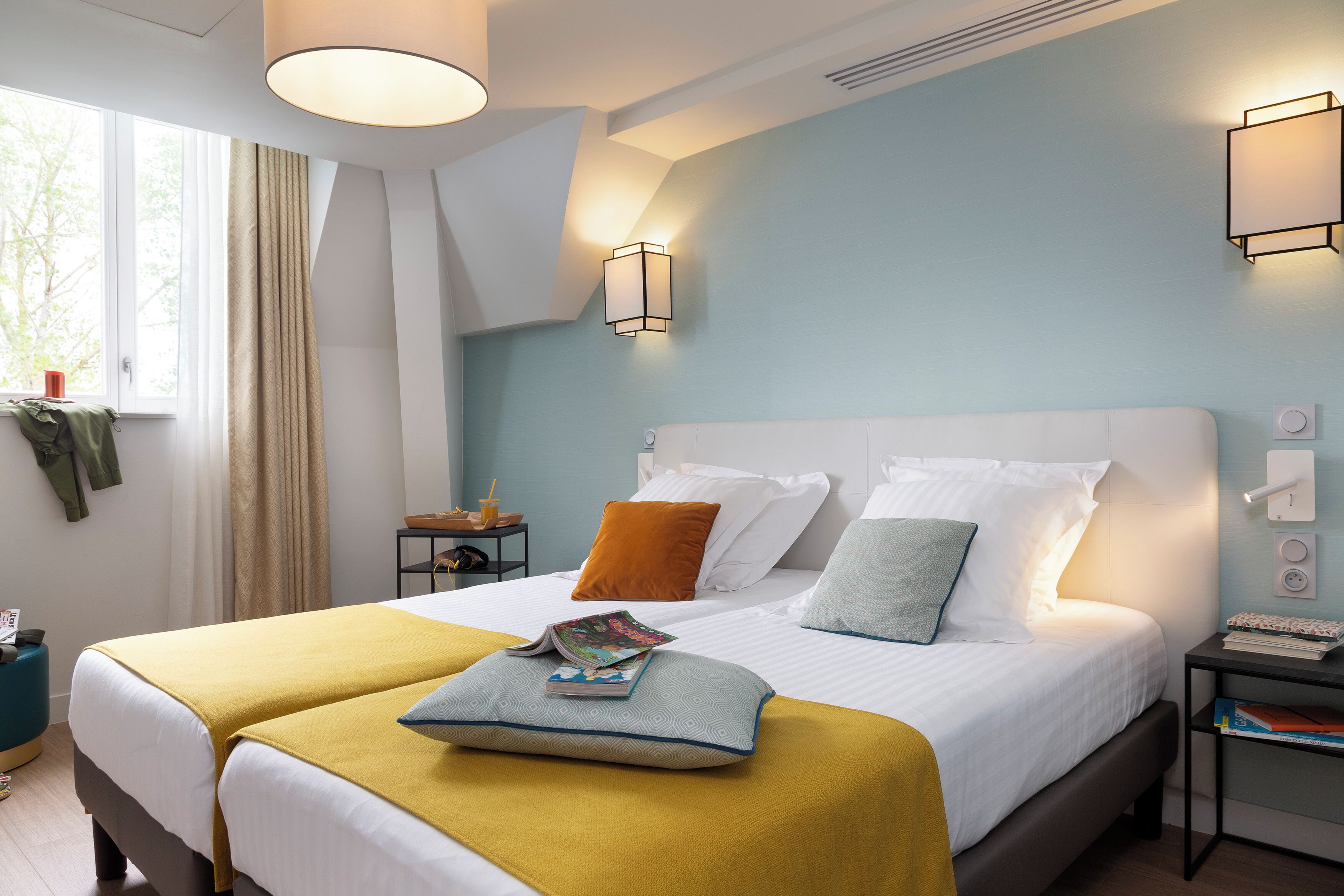 Une chambre à coucher avec un grand lit, deux tables de chevet, une fenêtre avec des rideaux et un luminaire fixé au mur.