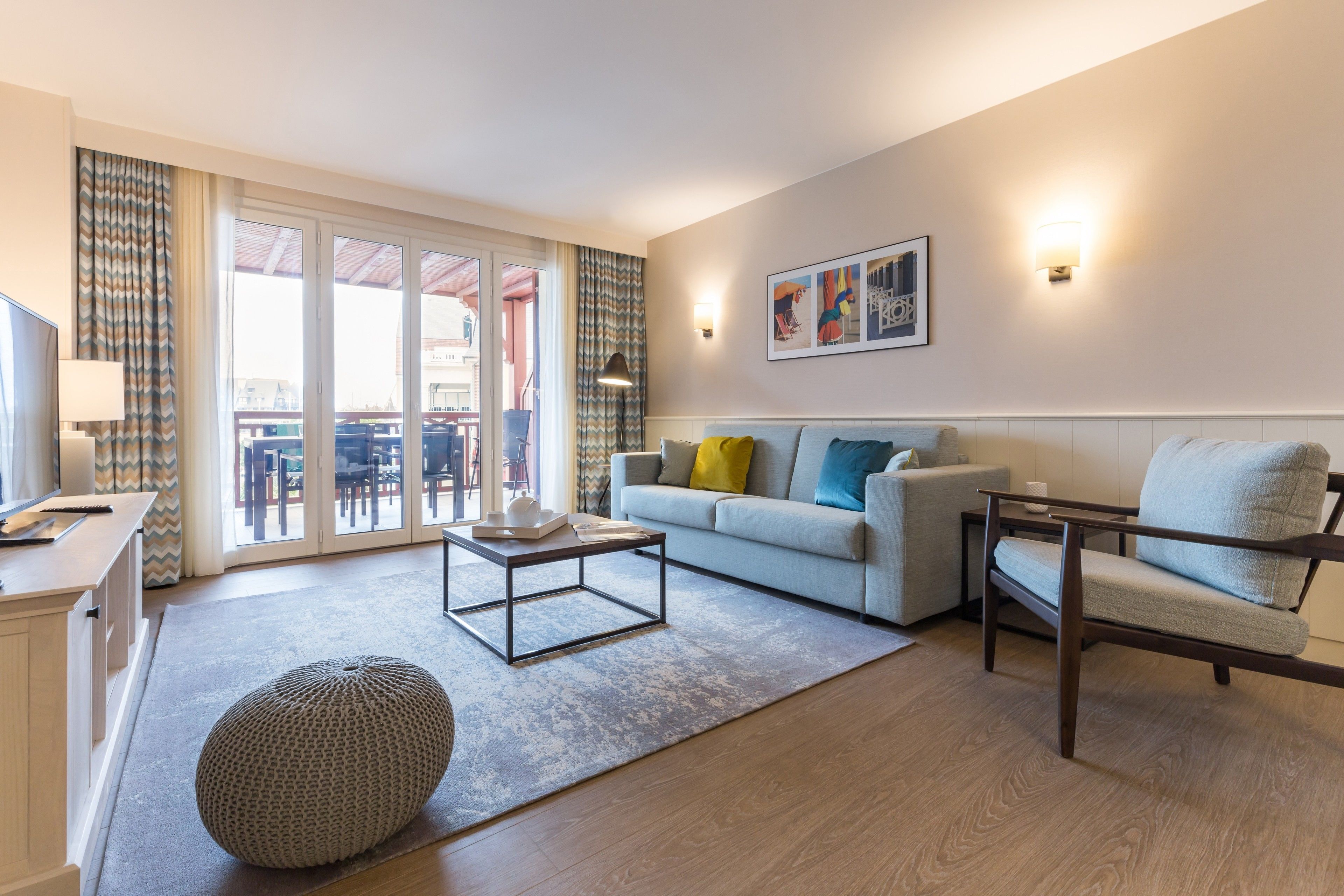 A modern living room with a sofa, armchair, ottoman, and coffee table. There is a balcony with a view of the city.