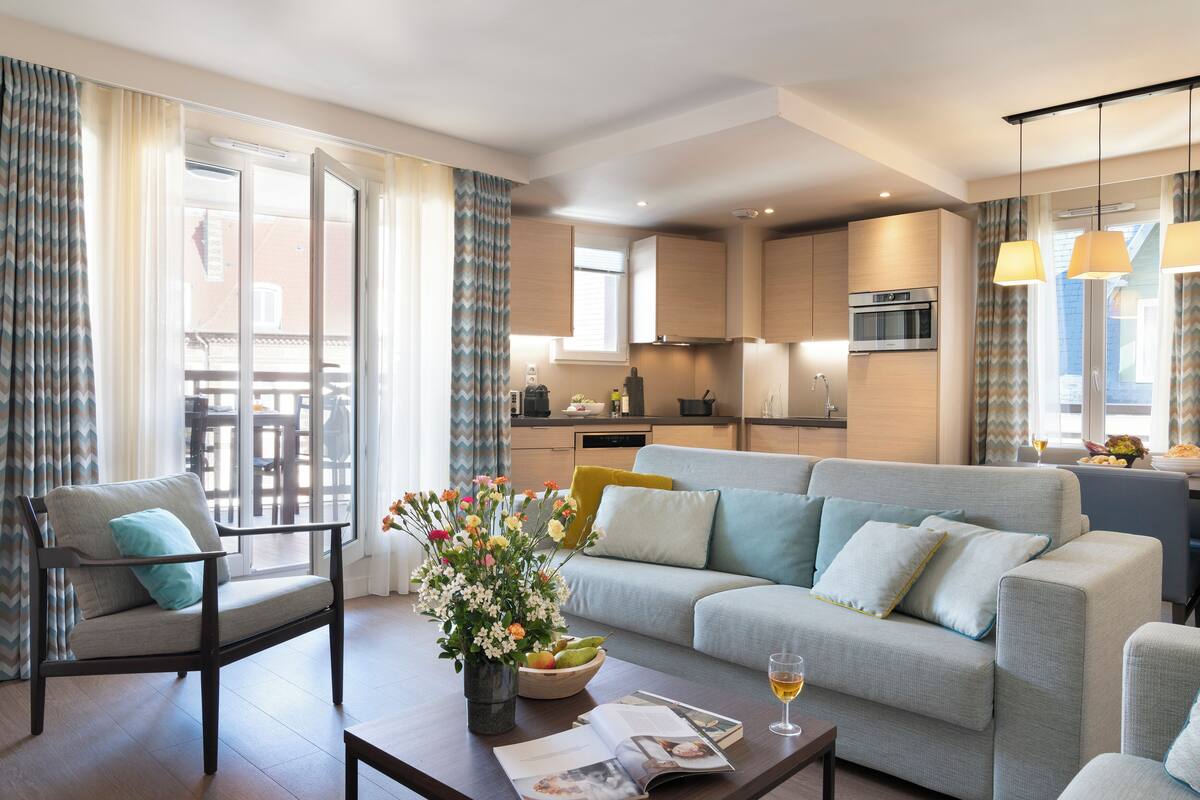 Uma sala de estar moderna com sofá, poltrona e mesa de centro, cozinha ao fundo e grandes janelas com cortinas.