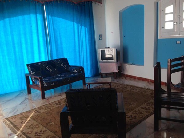 A living room with a blue sofa, a television on a stand, a wooden chair, and a patterned rug. - Luxor Guest House (Luxor)