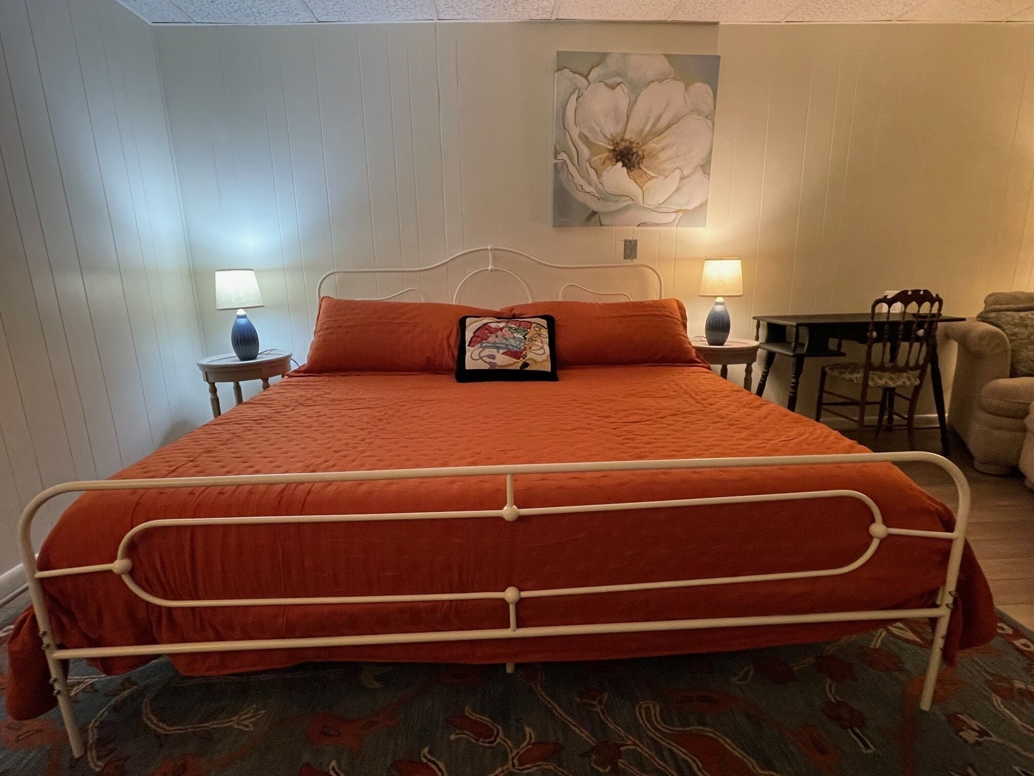 A bed with a metal frame, an orange bedspread, and a pillow with a floral design. There is a small table with a lamp and a chair next to the bed. A large flower painting is on the wall above the bed.