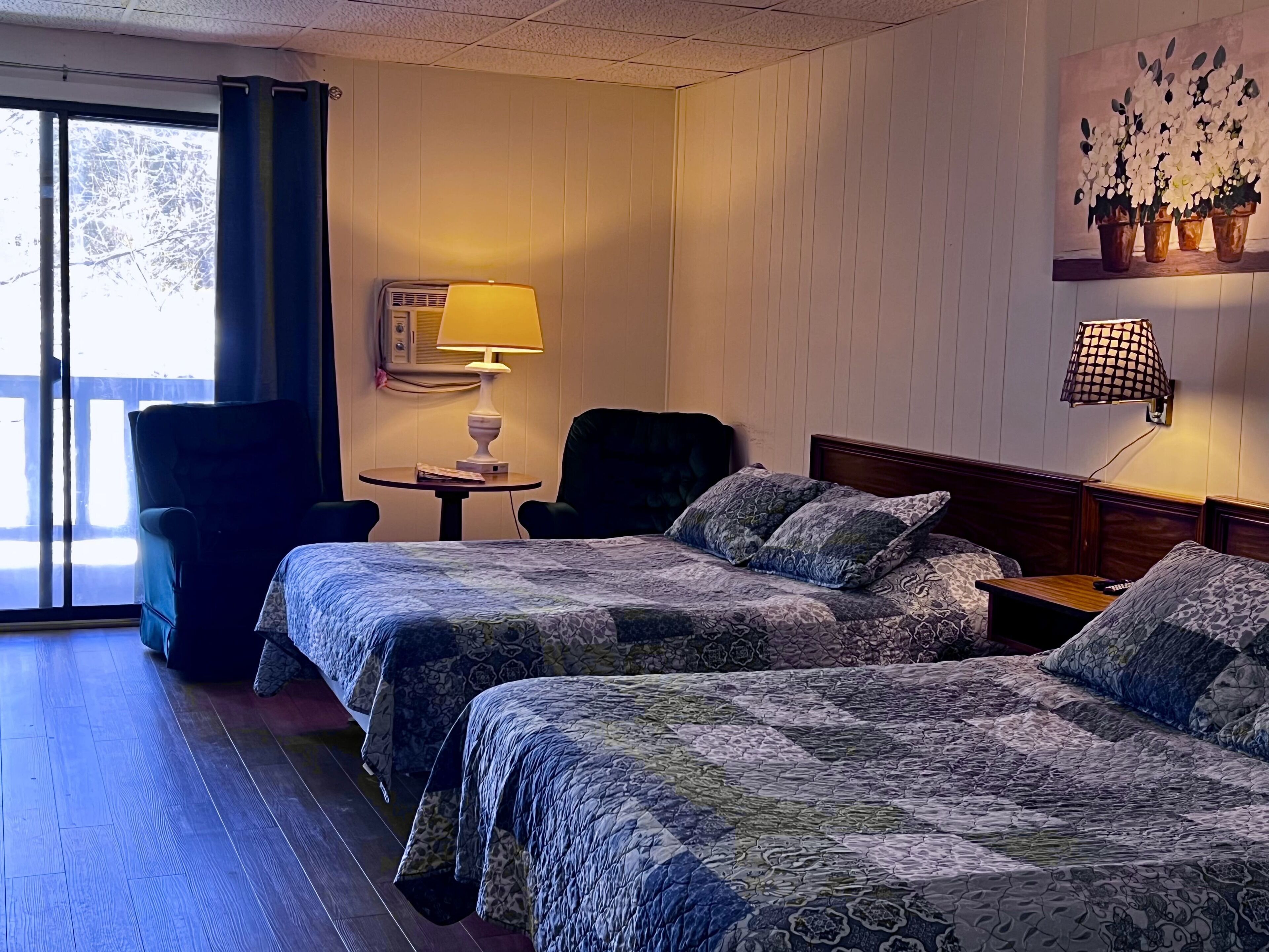 Habitación de hotel con dos camas, una silla, una mesita, una lámpara y un cuadro en la pared.