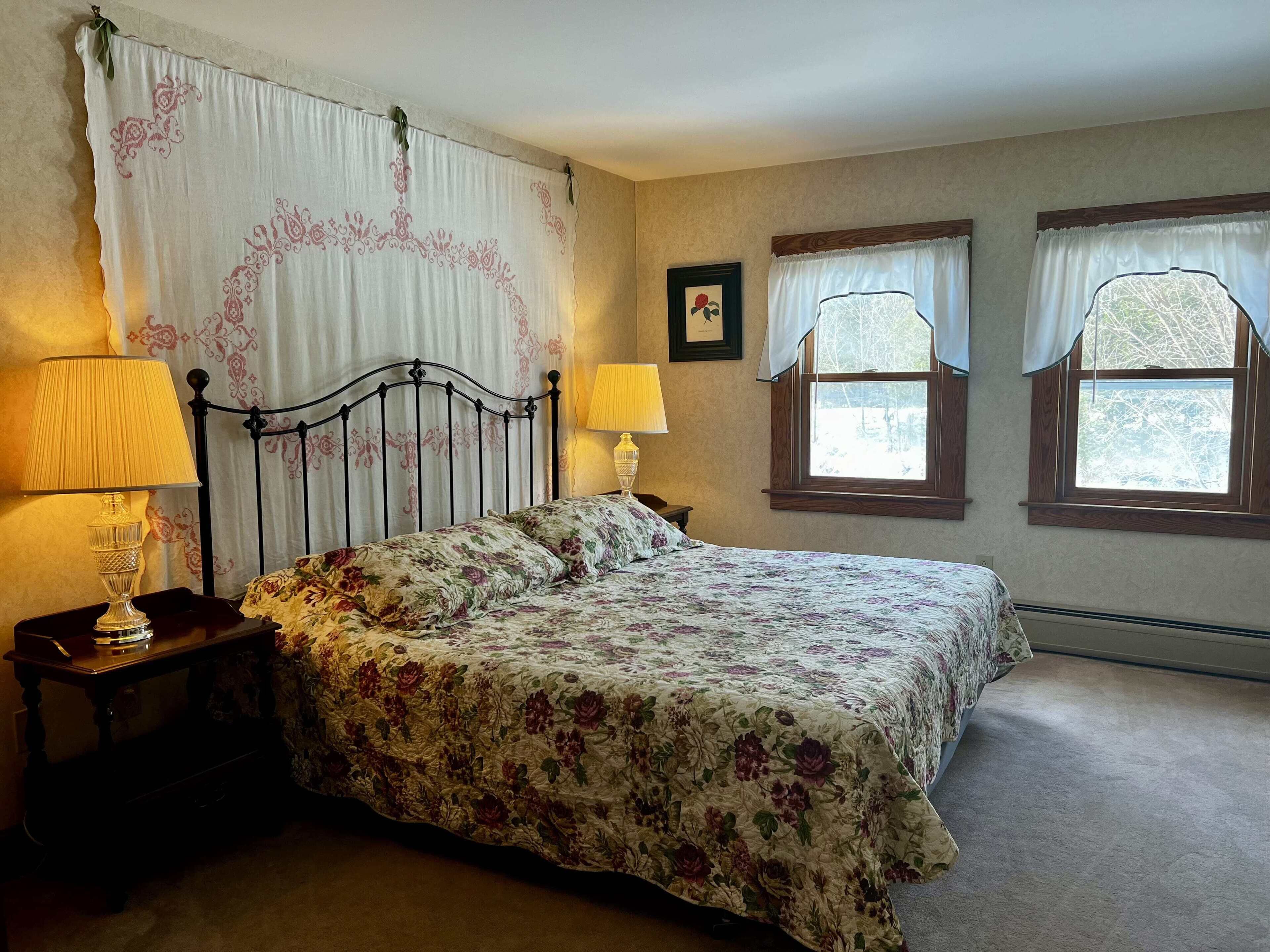 A bedroom with a bed, two bedside lamps, a nightstand, a window with curtains, and a framed picture on the wall.