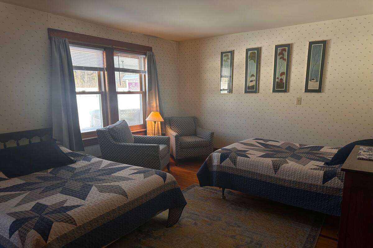 A hotel room with two beds, a chair, a lamp, and a window with curtains.