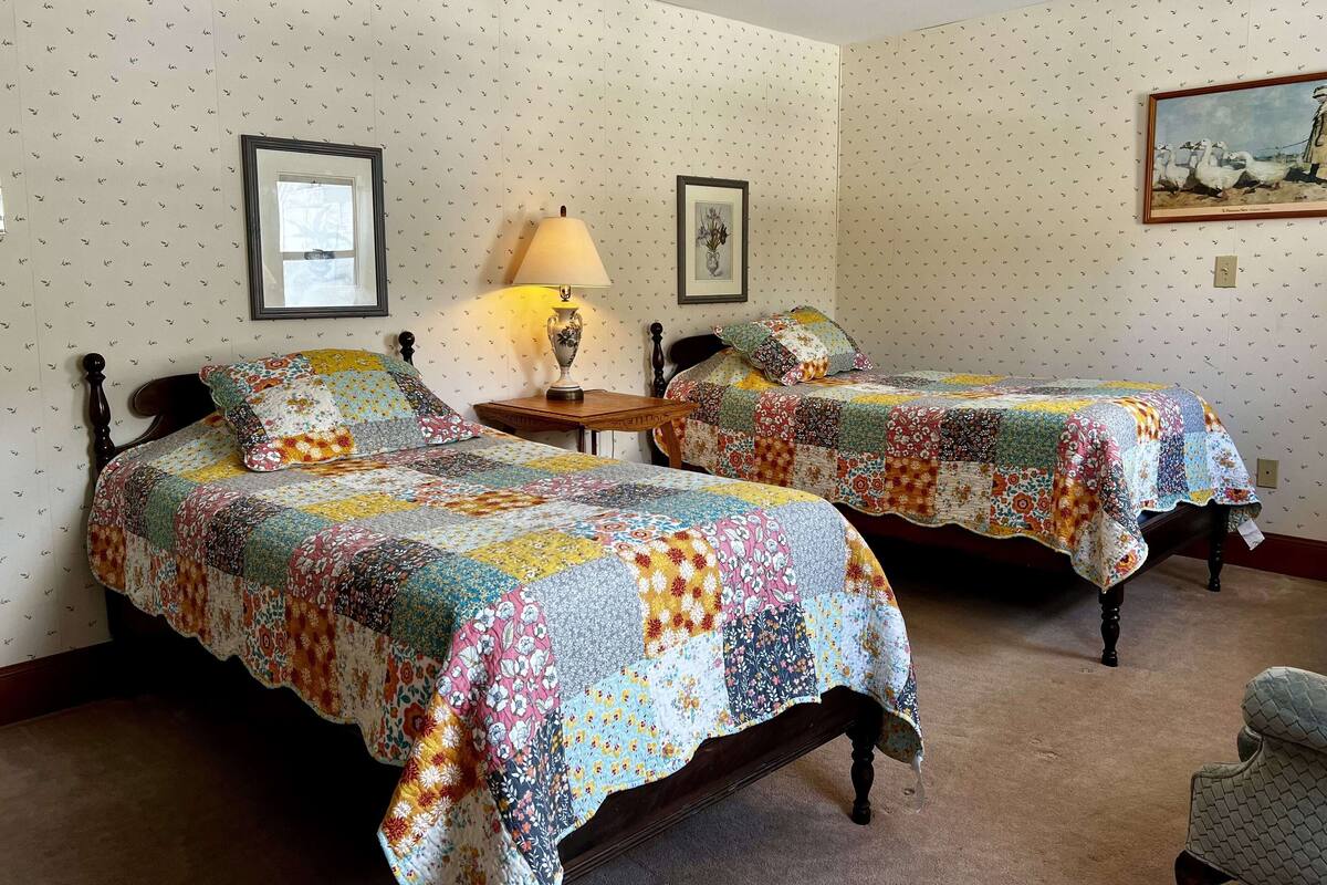 A room with two beds, each covered with a colorful quilt, a lamp on a wooden nightstand, and a framed picture on the wall.