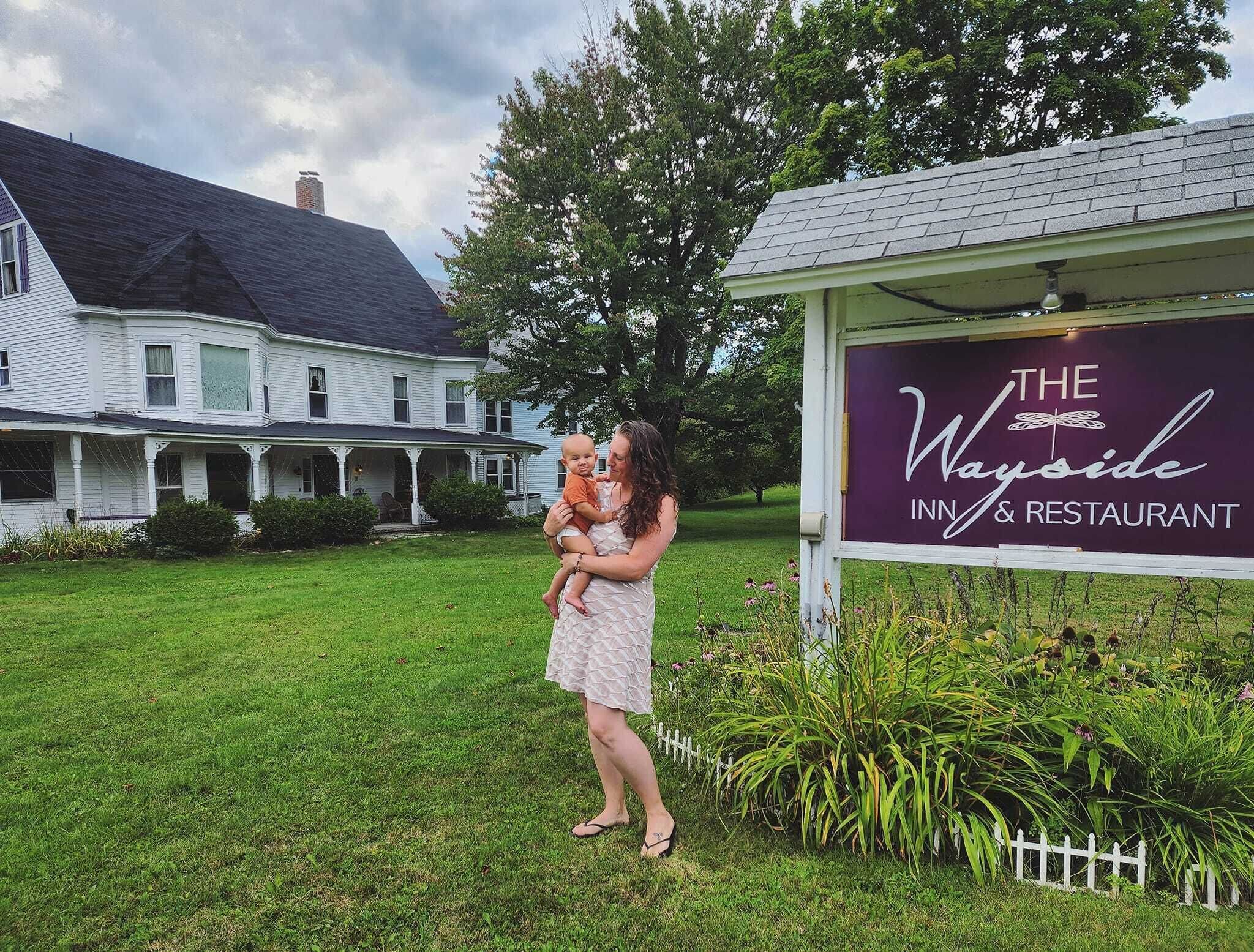 Una mujer sostiene a un bebé frente a una casa con un letrero que dice 'The Wayside Inn & Restaurant'.
