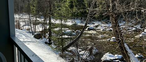A snowy river with rocks and trees, viewed from a balcony with a railing.