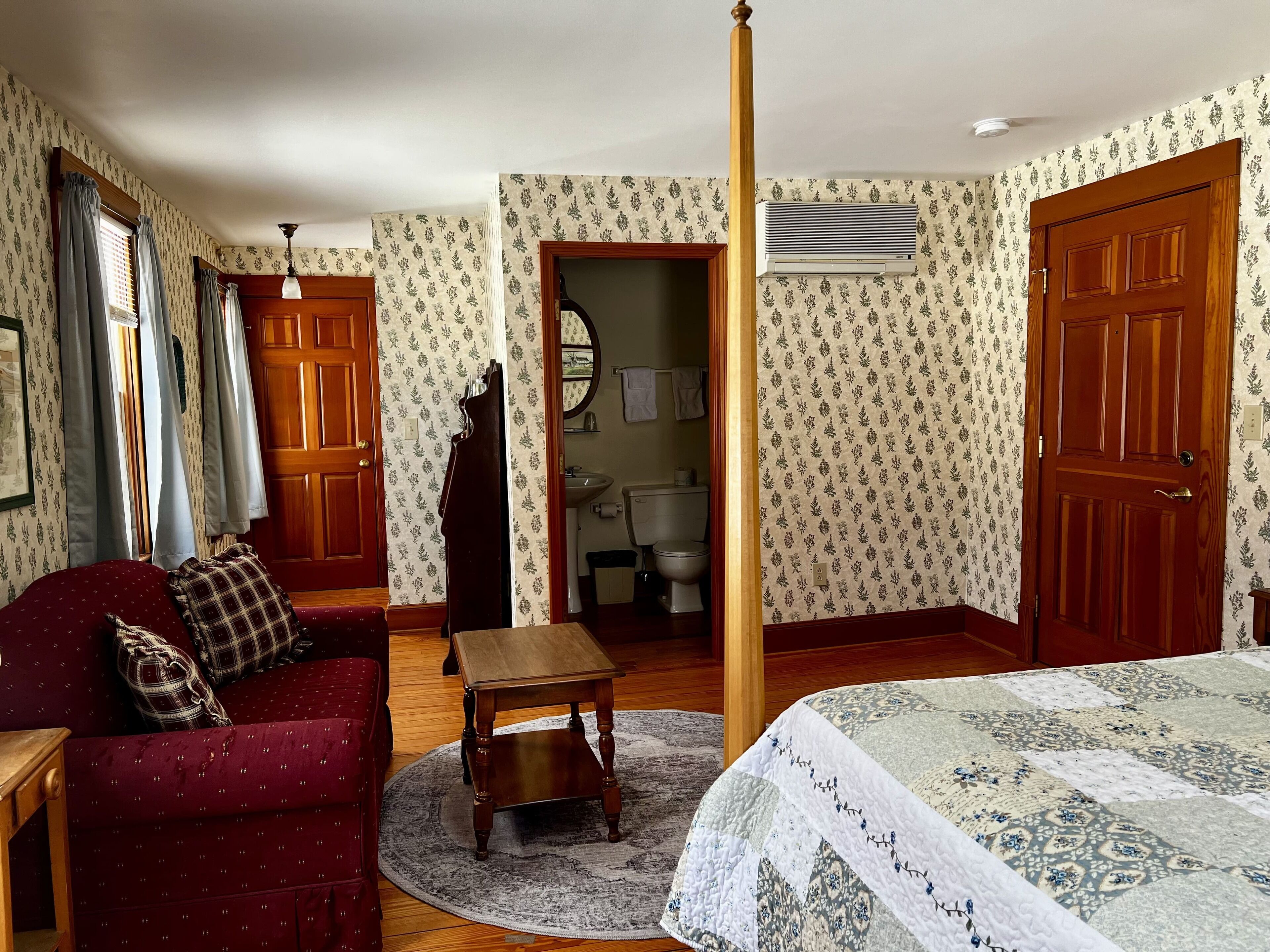 A room with a wooden floor, a red sofa with a plaid pillow, a wooden table, a round rug, a bed with a patterned quilt, a wooden door, a wooden wardrobe, a mirror, and a window with curtains.