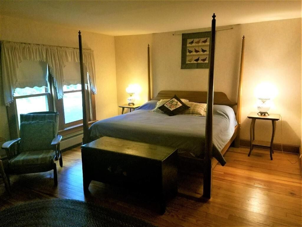 A four-poster bed with a nightstand on each side, a chair, and a small table in a warmly lit bedroom with wooden flooring.