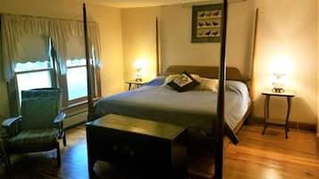A four-poster bed with a nightstand on each side, a chair, and a small table in a warmly lit bedroom with wooden flooring.