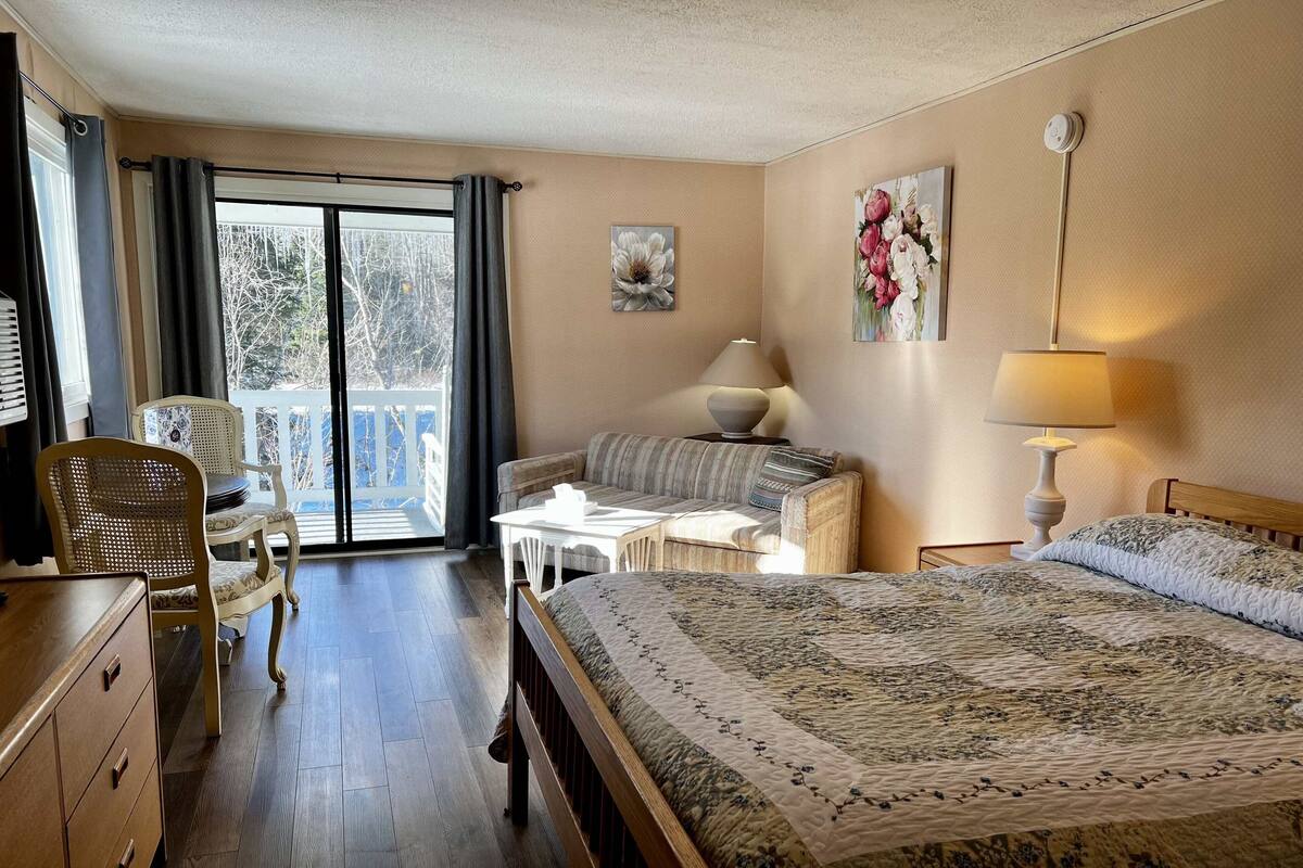 A hotel room with a bed, a sofa, a dining table, a chair, and a balcony with a view of trees.