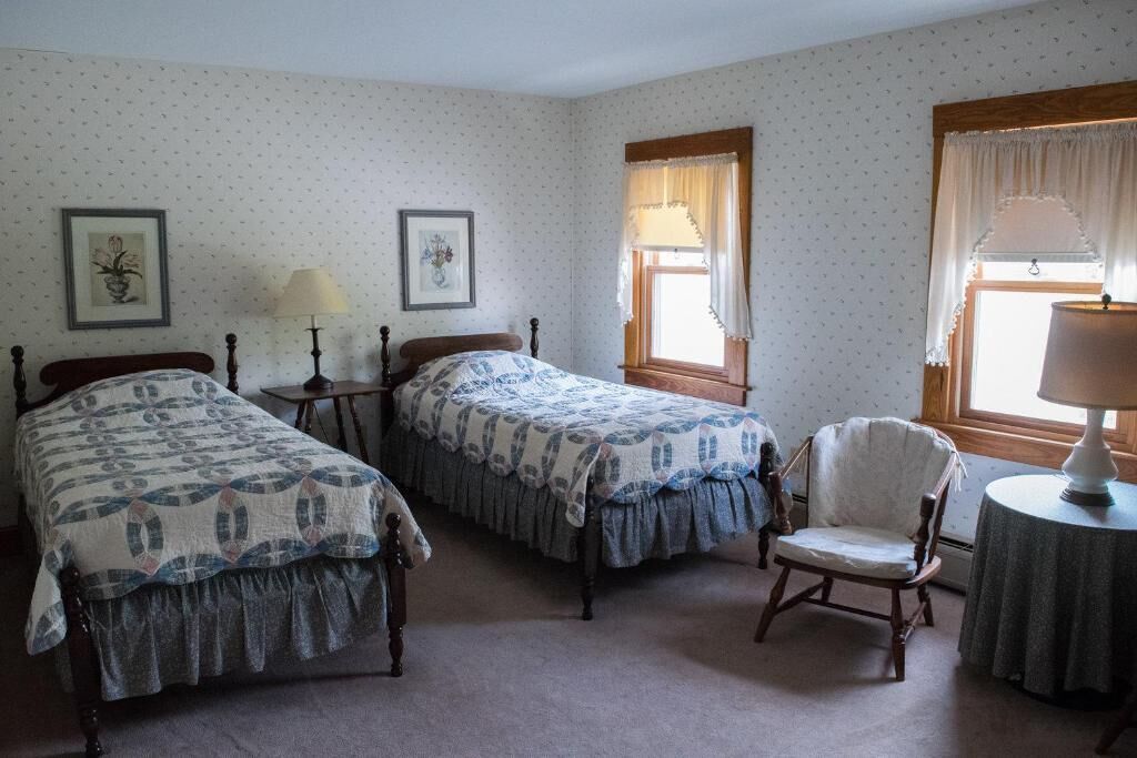 A room with two single beds, a chair, a table, and two windows with curtains.