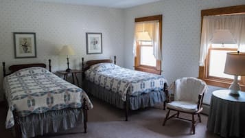 A room with two single beds, a chair, a table, and two windows with curtains.