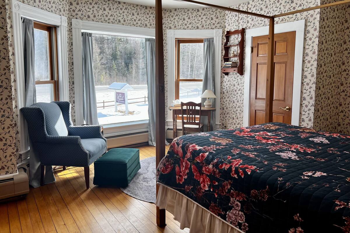 A bedroom with a four-poster bed, a chair, a desk, and a view of the outside.