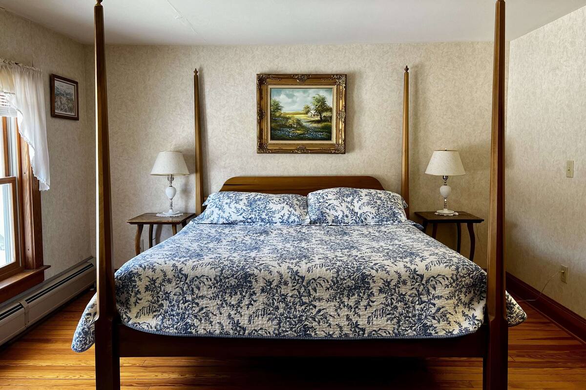 A four-poster bed with a floral bedspread, two bedside tables with lamps, a framed landscape painting on the wall, and a window with curtains.