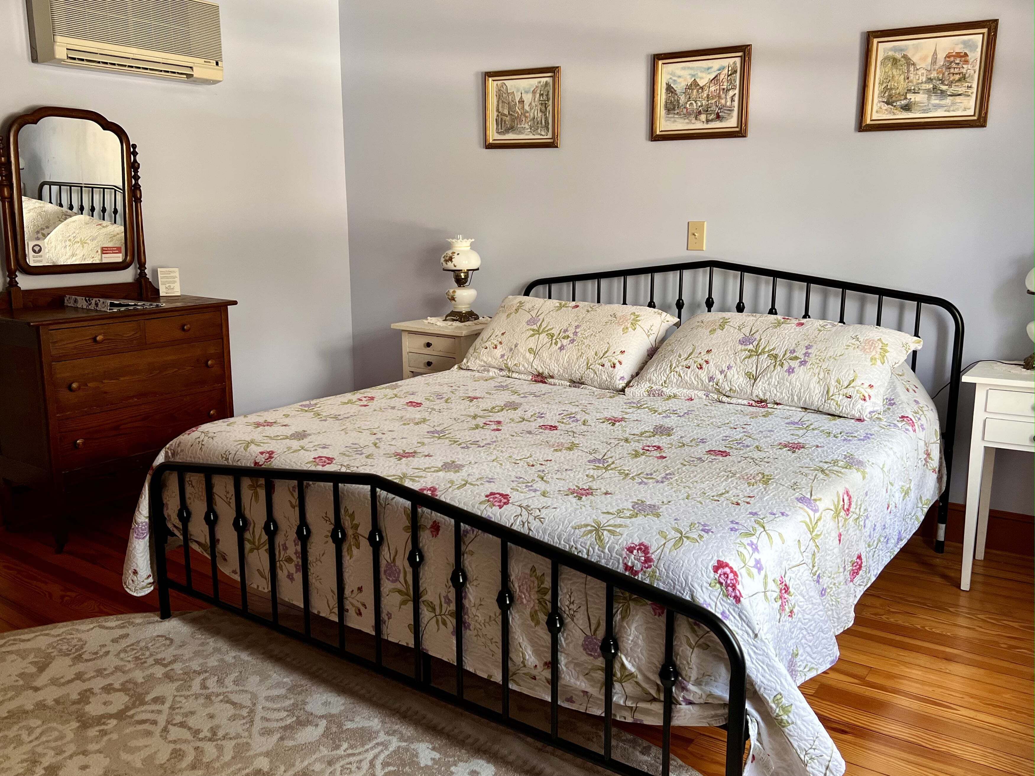 A bedroom with a floral bedspread, a wooden dresser, a nightstand with a lamp, and framed pictures on the wall.
