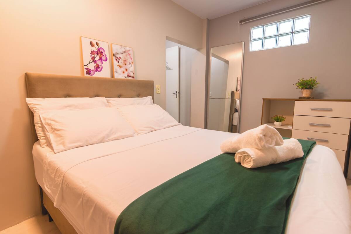Cama bem arrumada com lençóis brancos e manta verde, um pequeno vaso de planta em uma cômoda, e uma janela com luz natural.
