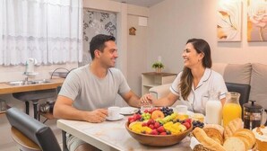A dining area with a table set for two, a bowl of fruit, and a breakfast spread including pastries and beverages. - Flores de Bombinhas (Bombinhas)