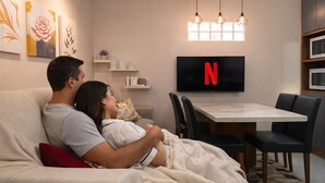 A couple on a sofa with a TV displaying the Netflix logo, a dining area with chairs and a table, and artwork on the walls. - Flores de Bombinhas (Bombinhas)
