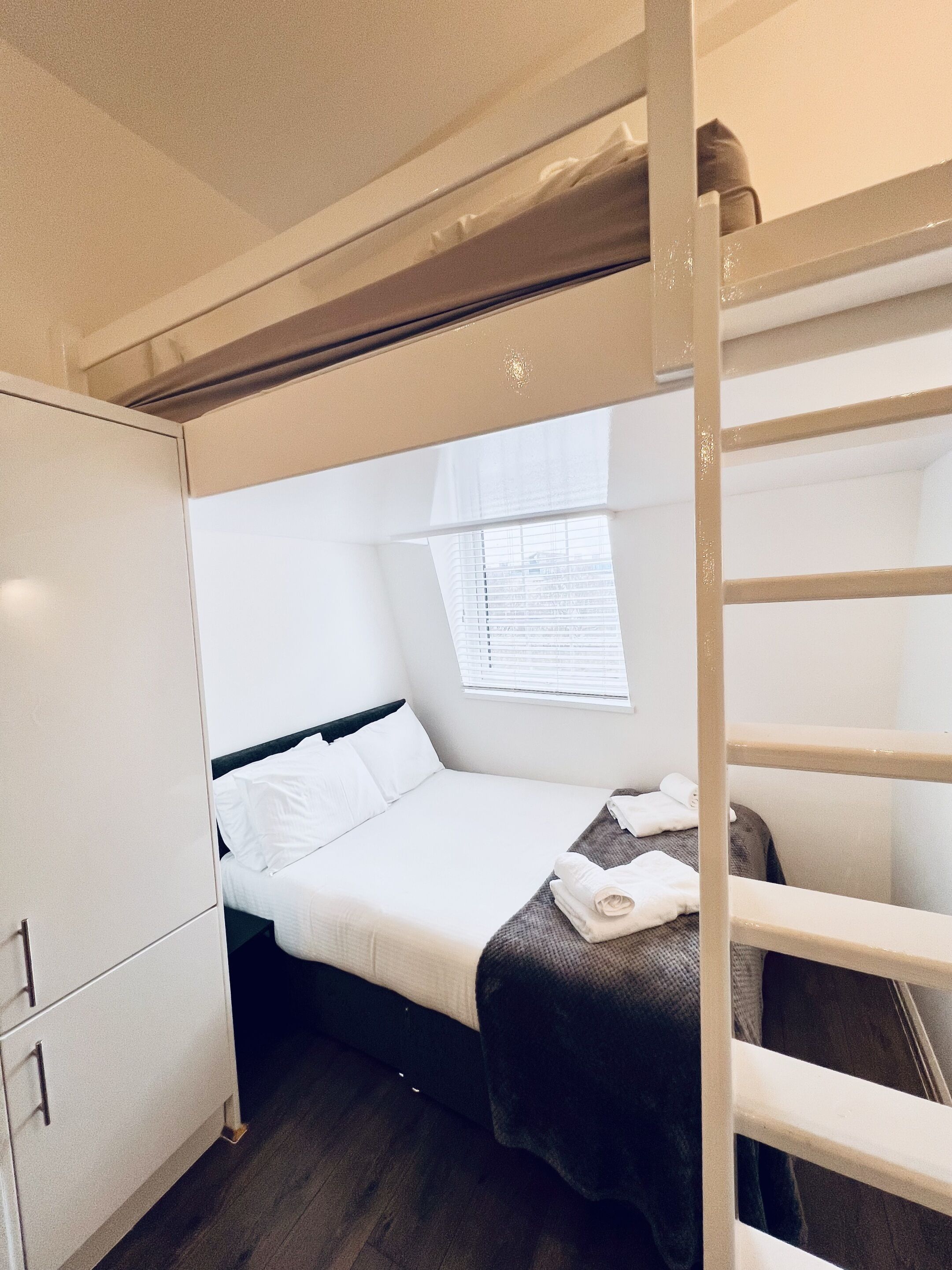 A compact room with a bunk bed, a small window with blinds, a wardrobe, and a neatly made bed with a blanket and pillows.
