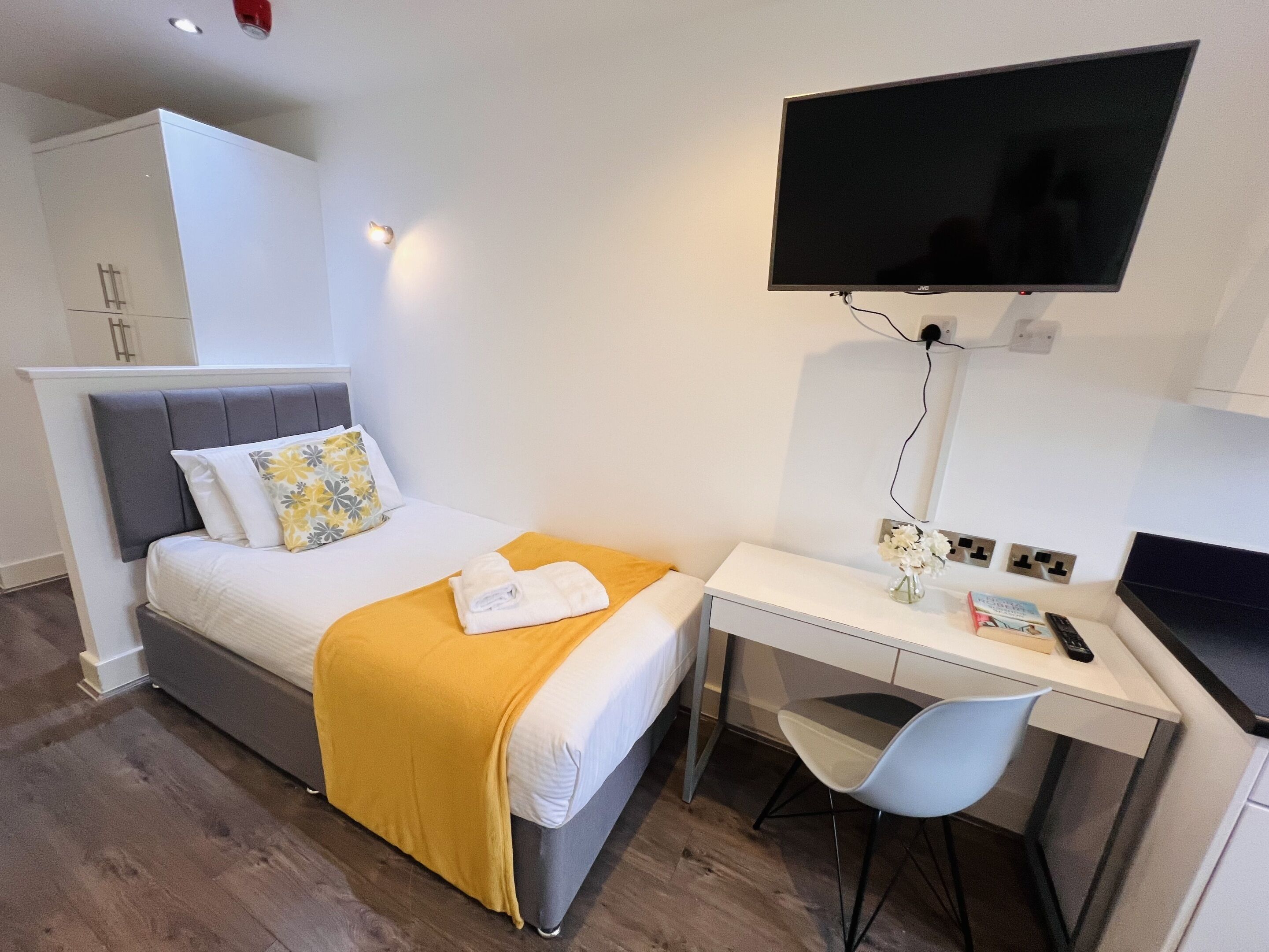 Une chambre d’hôtel avec un lit, un bureau, une chaise, une télévision et une petite table sur laquelle se trouvent un vase et des livres.