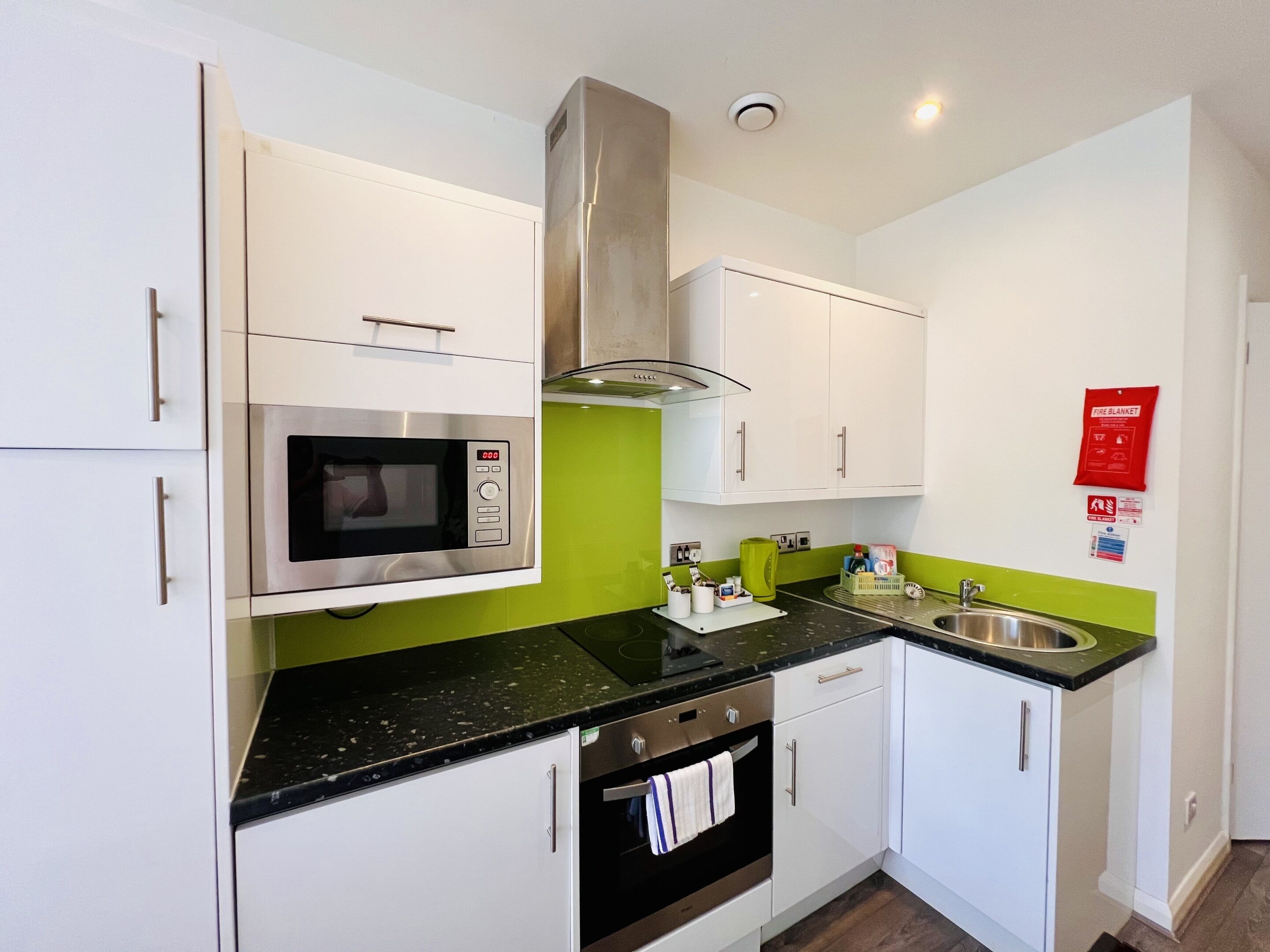 Una cocina compacta con armarios blancos, un microondas de acero inoxidable, horno empotrado y salpicadero verde.