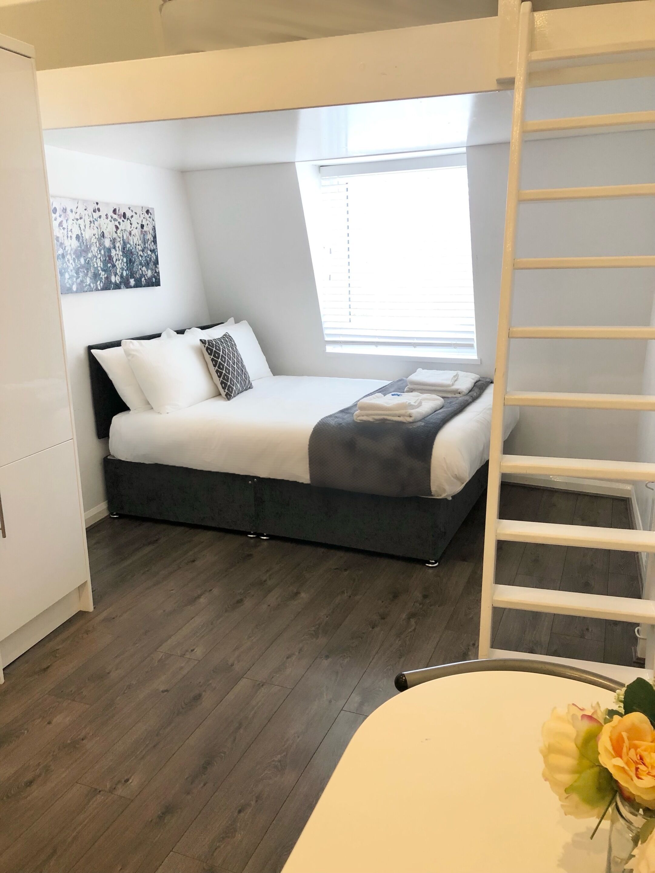 Une petite chambre à coucher comprenant un lit, une fenêtre, une échelle et une petite table sur laquelle est posé un vase de fleurs.