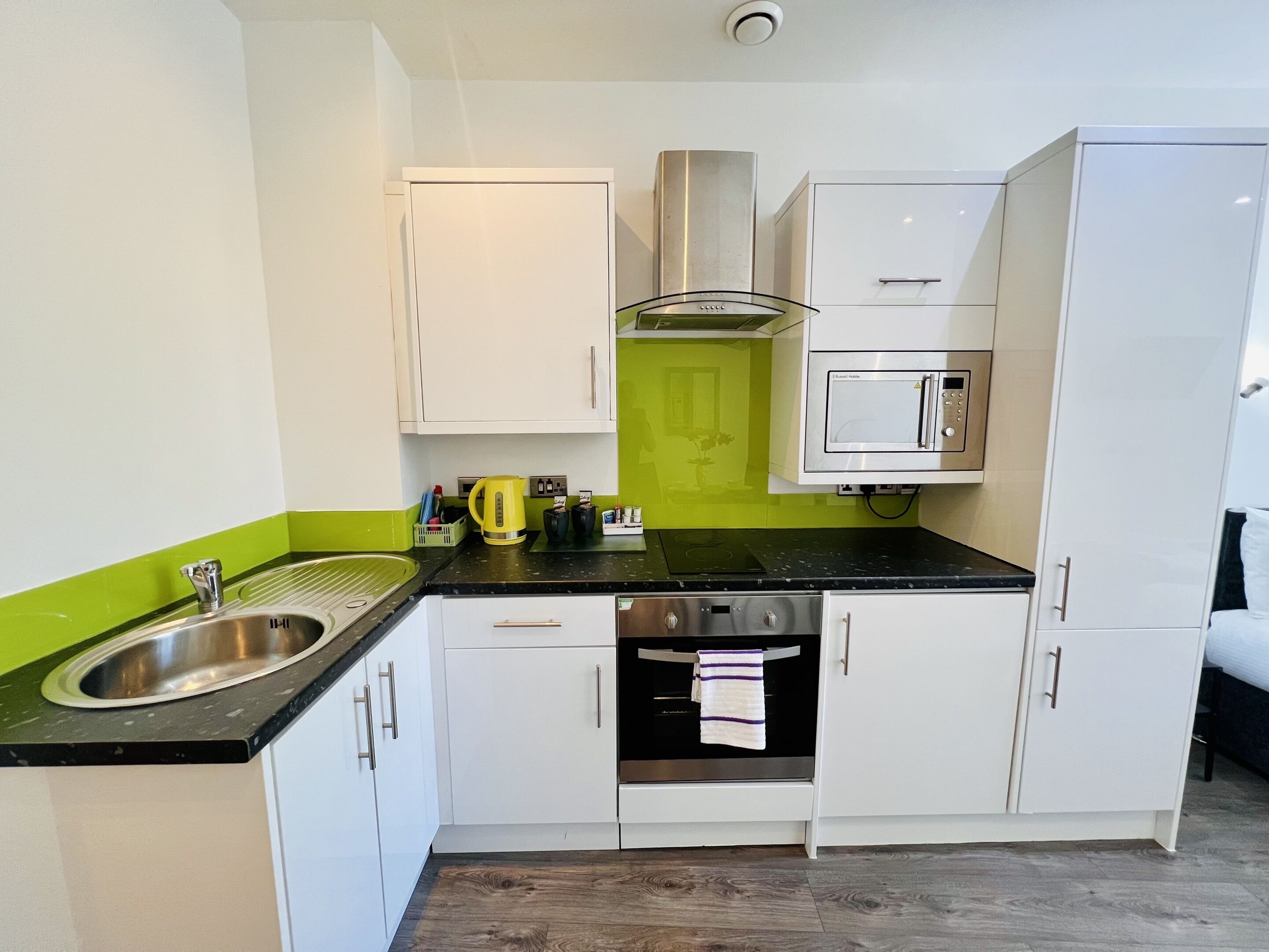 A compact kitchen with white cabinets, a black countertop, a stainless steel sink, a kettle, a microwave, and a stove.