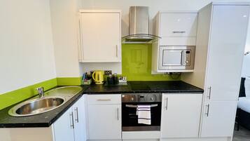 A compact kitchen with white cabinets, a black countertop, a stainless steel sink, a kettle, a microwave, and a stove.