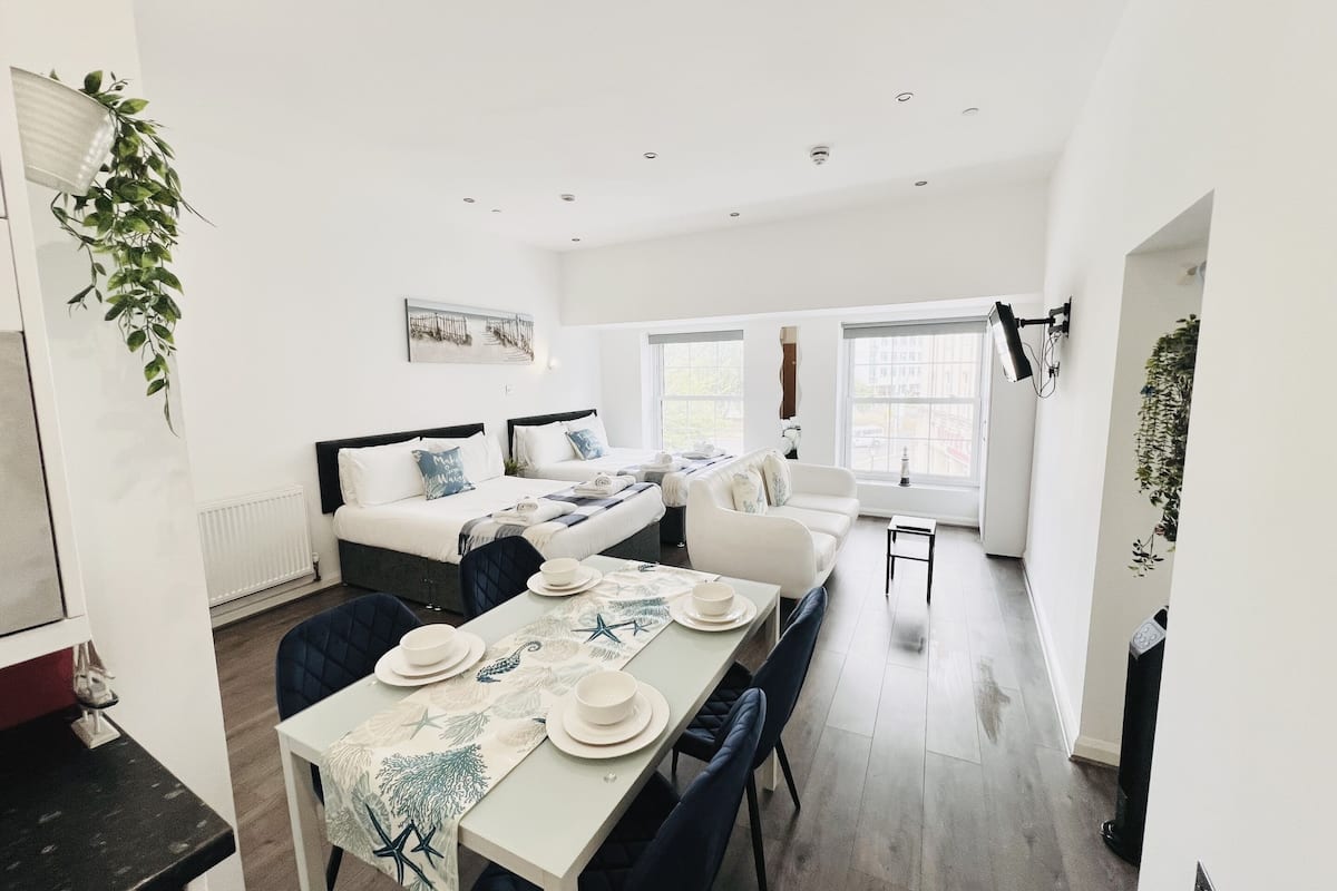 A modern, well-lit room with a dining table set for four, two beds, and a large window providing natural light.