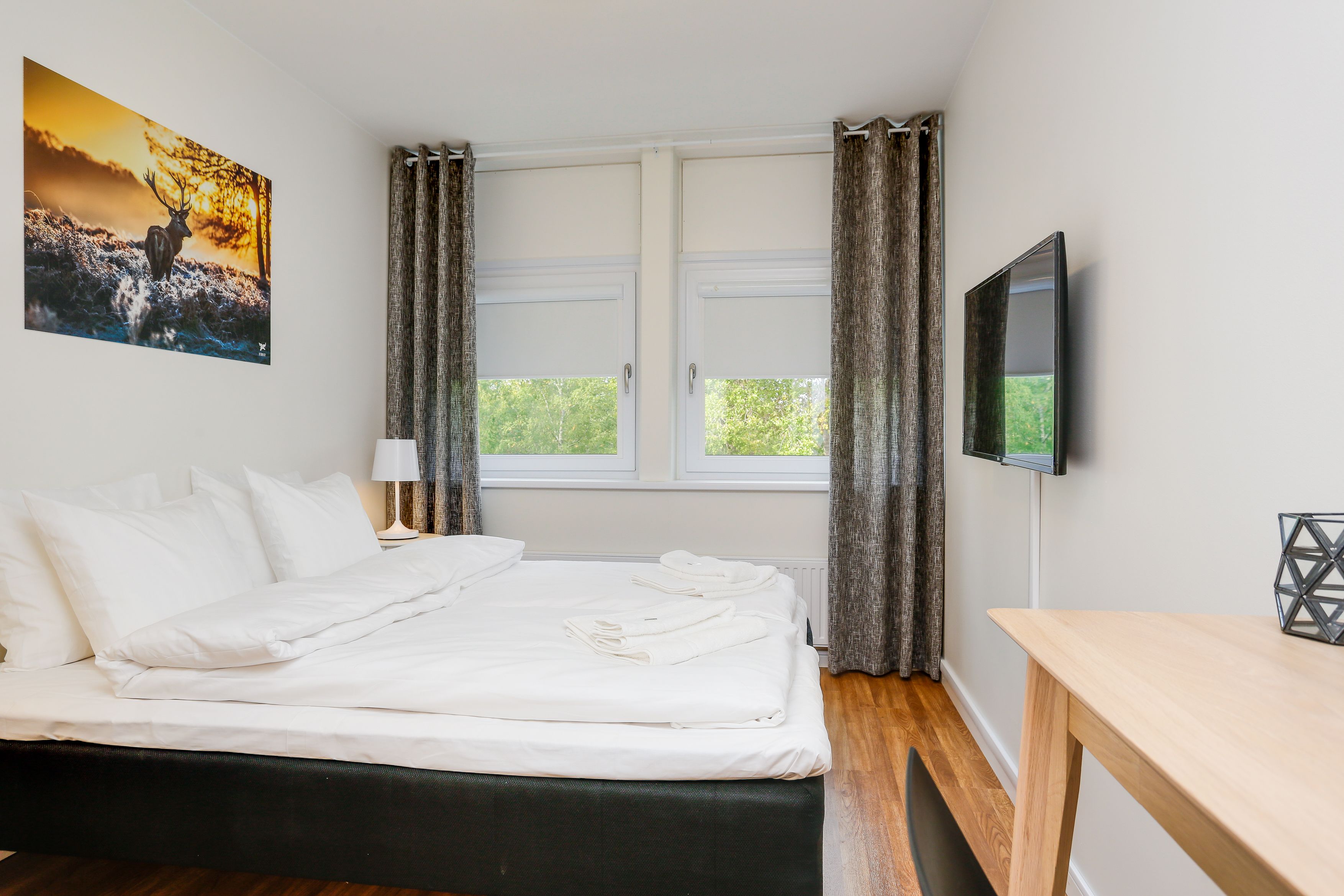Una cama bien hecha con ropa blanca, una mesita de noche con lámpara, un televisor en la pared y una ventana con cortinas.