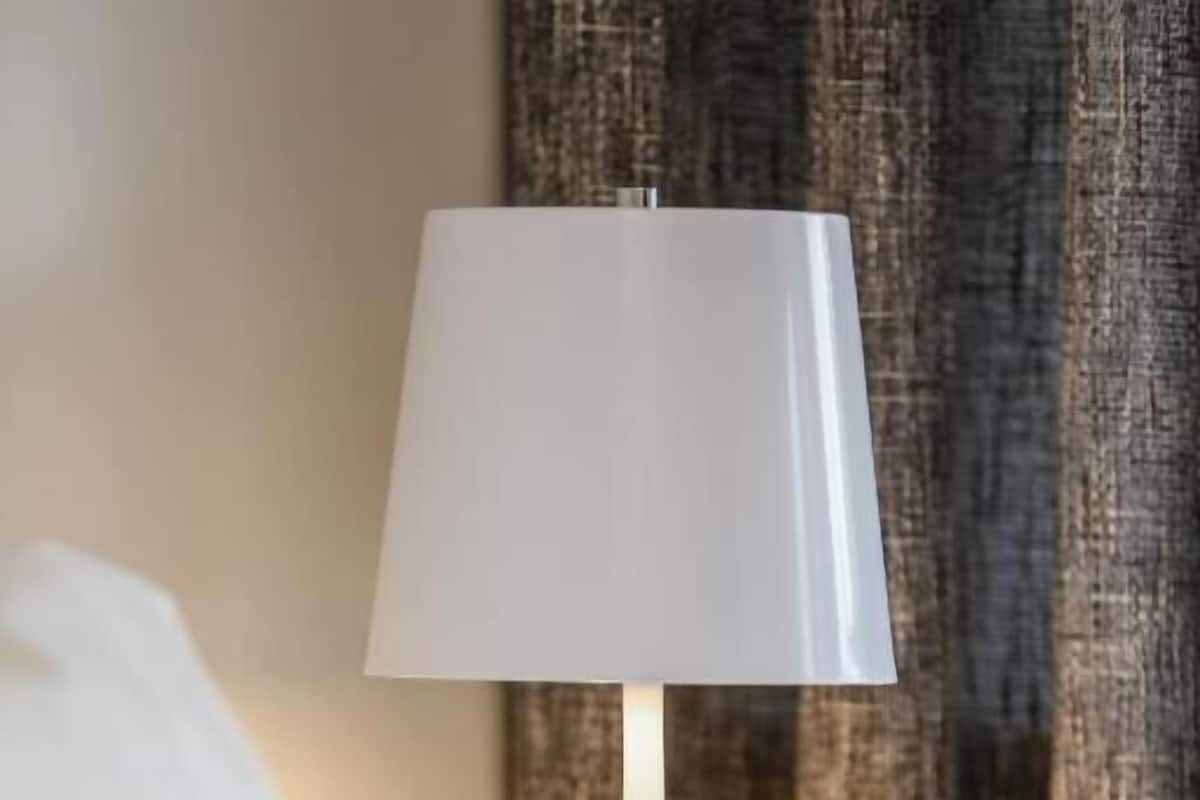 Une lampe de chevet blanche, posée sur une table de nuit, avec un rideau à motifs en arrière-plan.