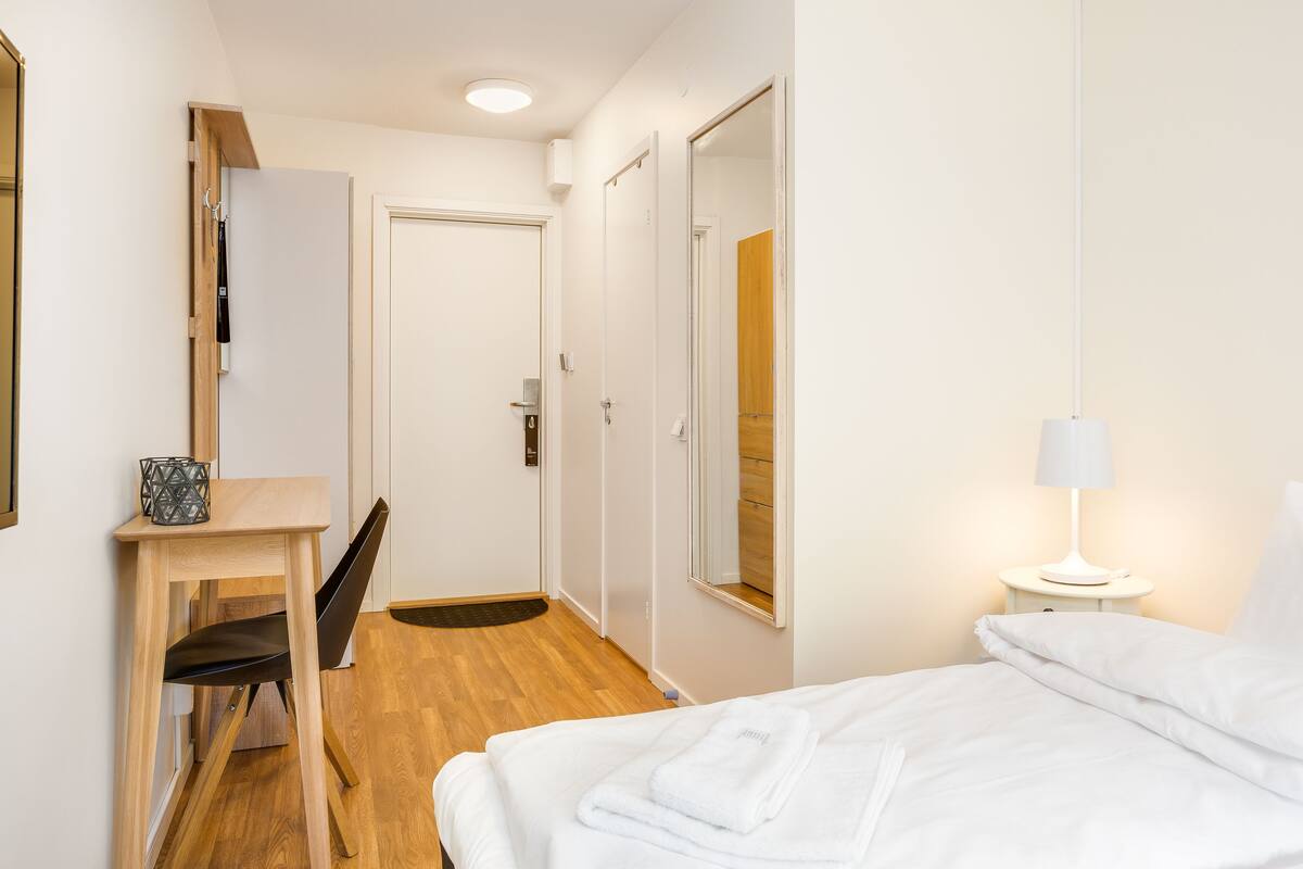 Une chambre d’hôtel comprenant un lit, un bureau avec une chaise, un miroir, une table de chevet avec une lampe et une porte donnant sur une autre pièce.