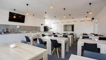 A modern dining area with multiple tables and chairs, pendant lighting, and a kitchen area in the background.