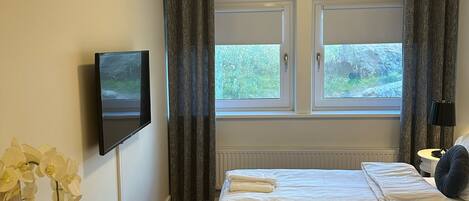A hotel room with a bed, a TV, a window with curtains, a radiator, and a small table with a vase of flowers.