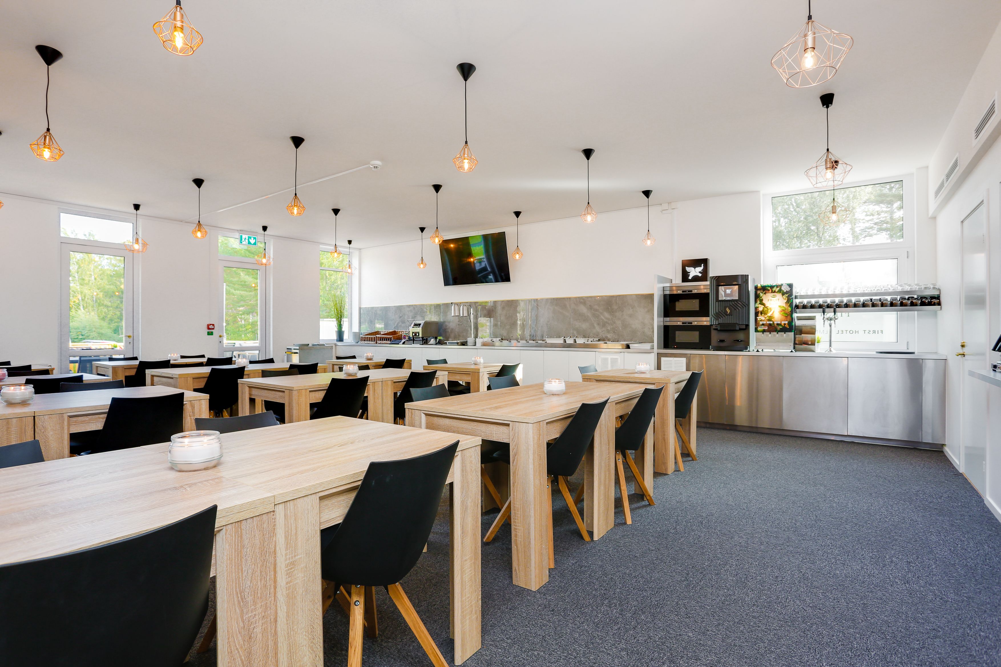 Uma sala de jantar moderna com mesas de madeira e cadeiras pretas, uma área de cozinha com eletrodomésticos de aço inoxidável e iluminação pendente.