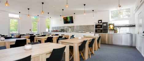 Uma sala de jantar moderna com mesas de madeira e cadeiras pretas, uma área de cozinha com eletrodomésticos de aço inoxidável e iluminação pendente.
