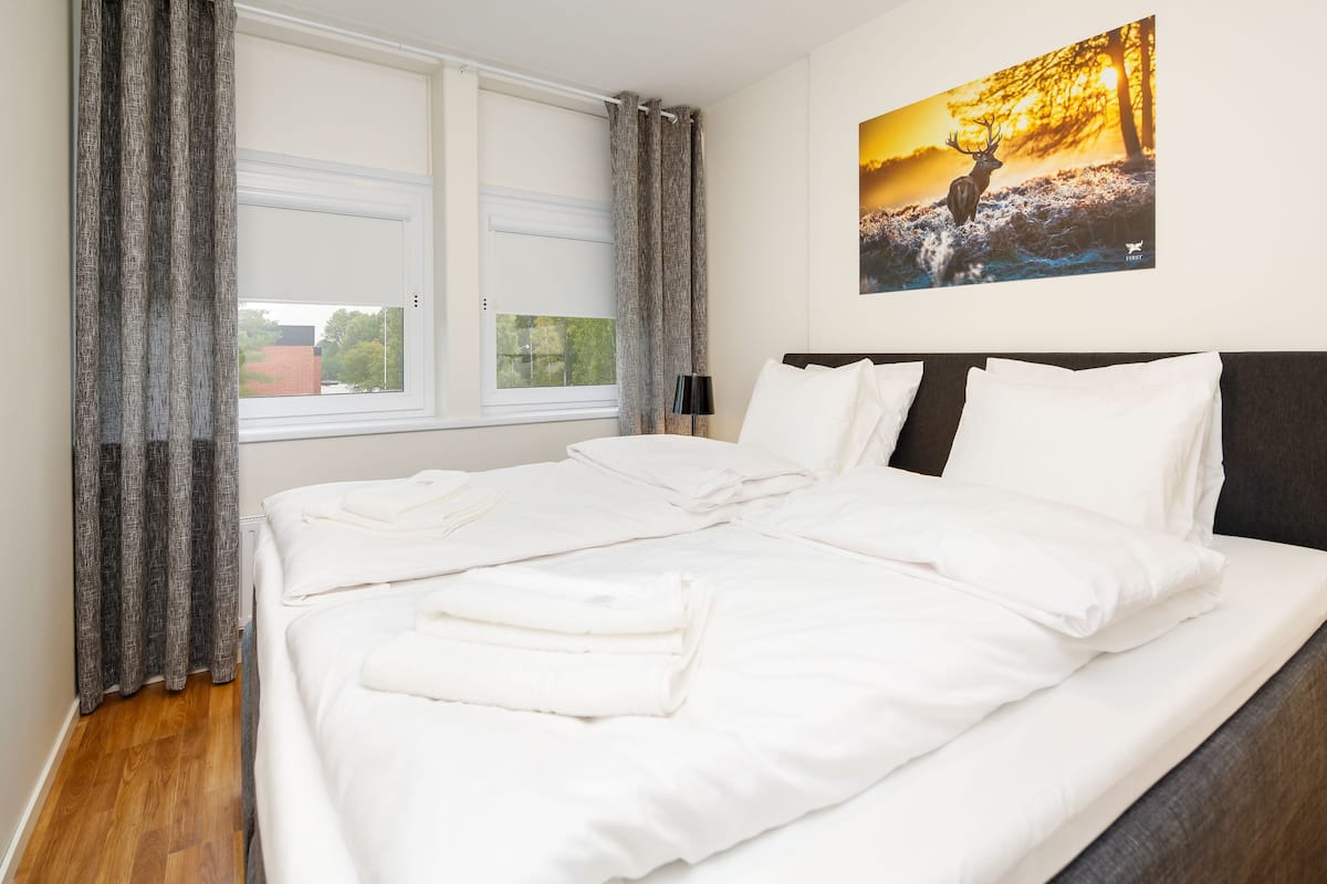 Un lit bien fait, avec du linge de lit blanc et des oreillers, une fenêtre avec des rideaux et un tableau représentant un cerf accroché au mur.