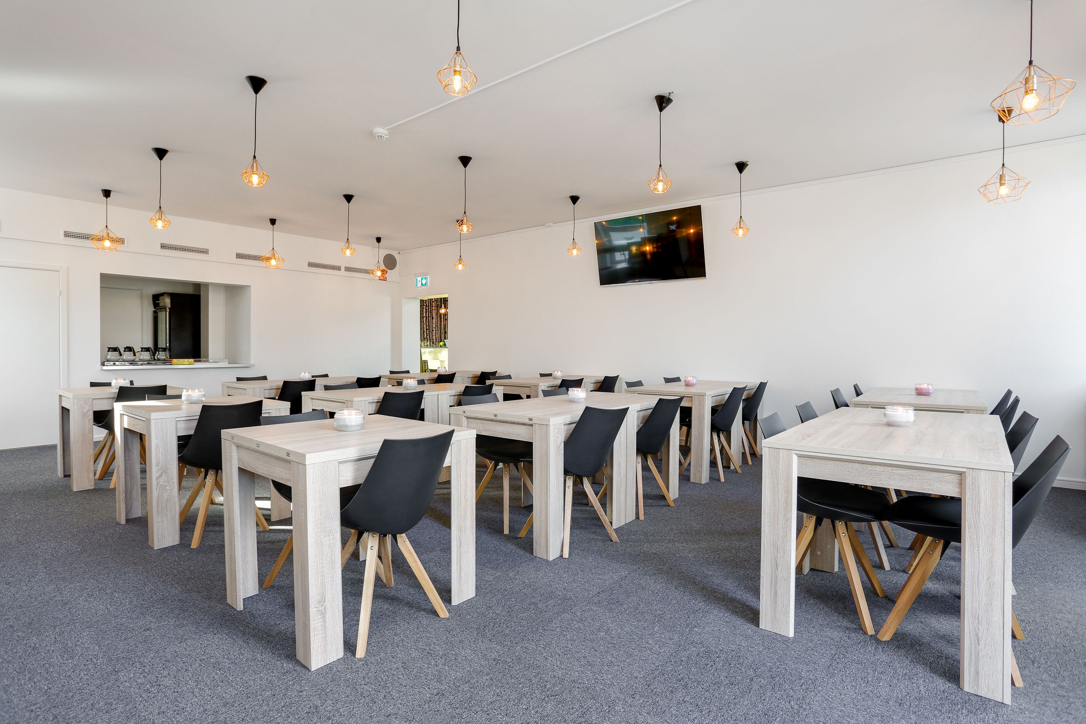A conference room with multiple tables and chairs, overhead lighting, and a mounted screen on the wall.