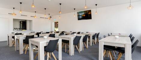 A conference room with multiple tables and chairs, overhead lighting, and a mounted screen on the wall.
