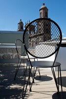 A chair with a metal mesh backrest and another chair with a solid backrest on a balcony with a clear view of a building with domed towers.