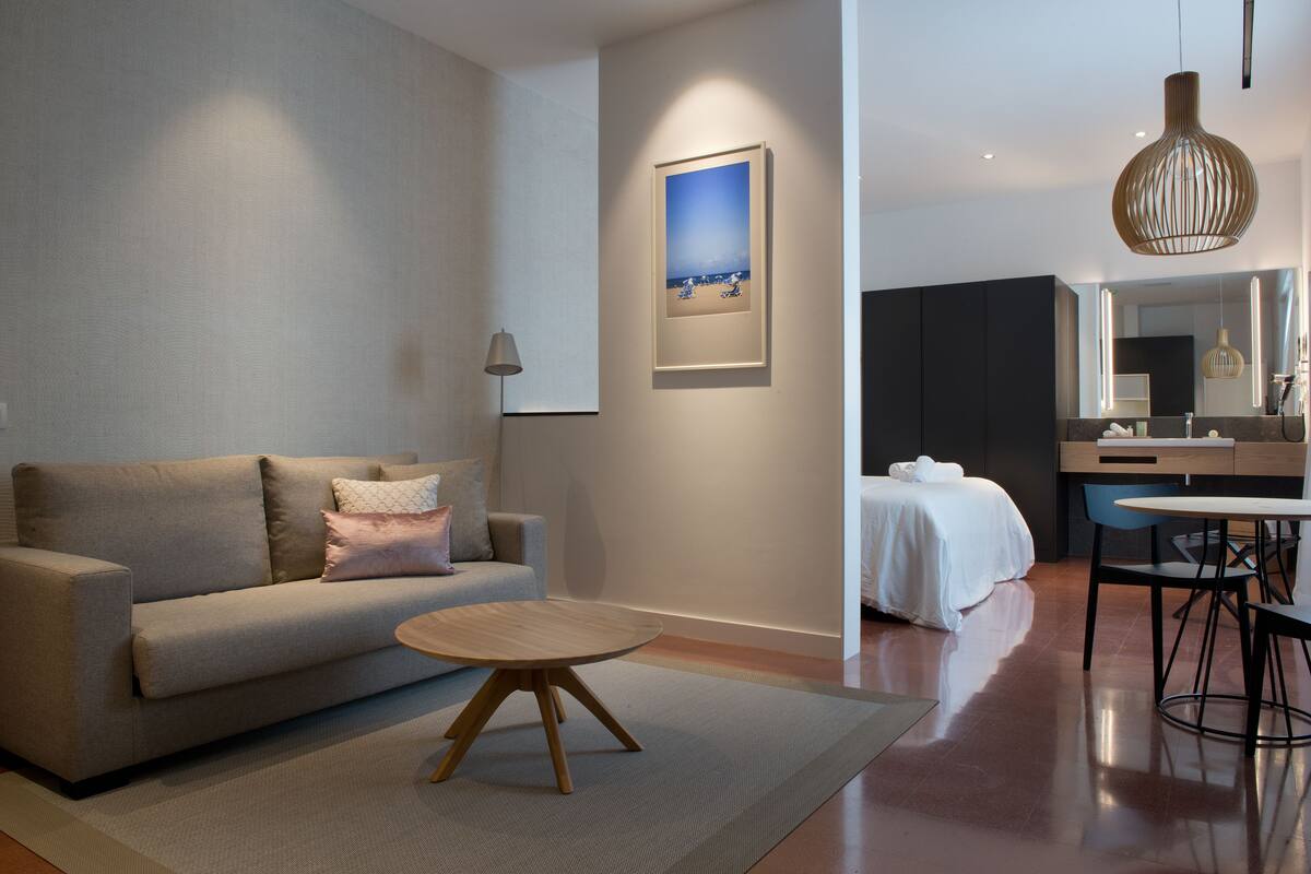 Ein modernes Hotelzimmer mit einer Couch, einem runden Holztisch, einem Bett mit weißer Bettdecke, einem kleinen Schreibtisch mit Stuhl und einem großen Spiegel.