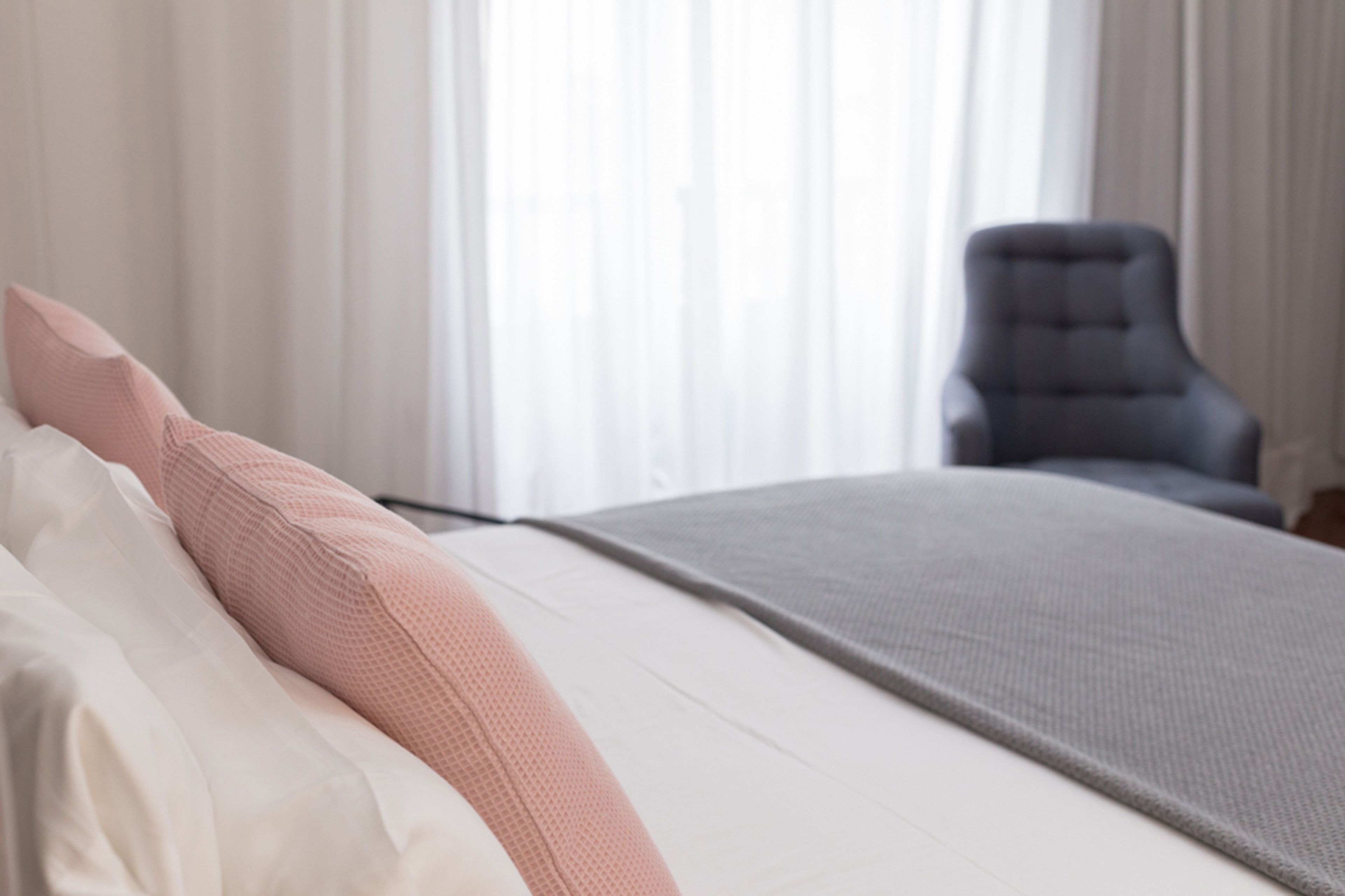 Una cama bien hecha con colcha blanca y gris, una almohada rosa y una silla gris al fondo.