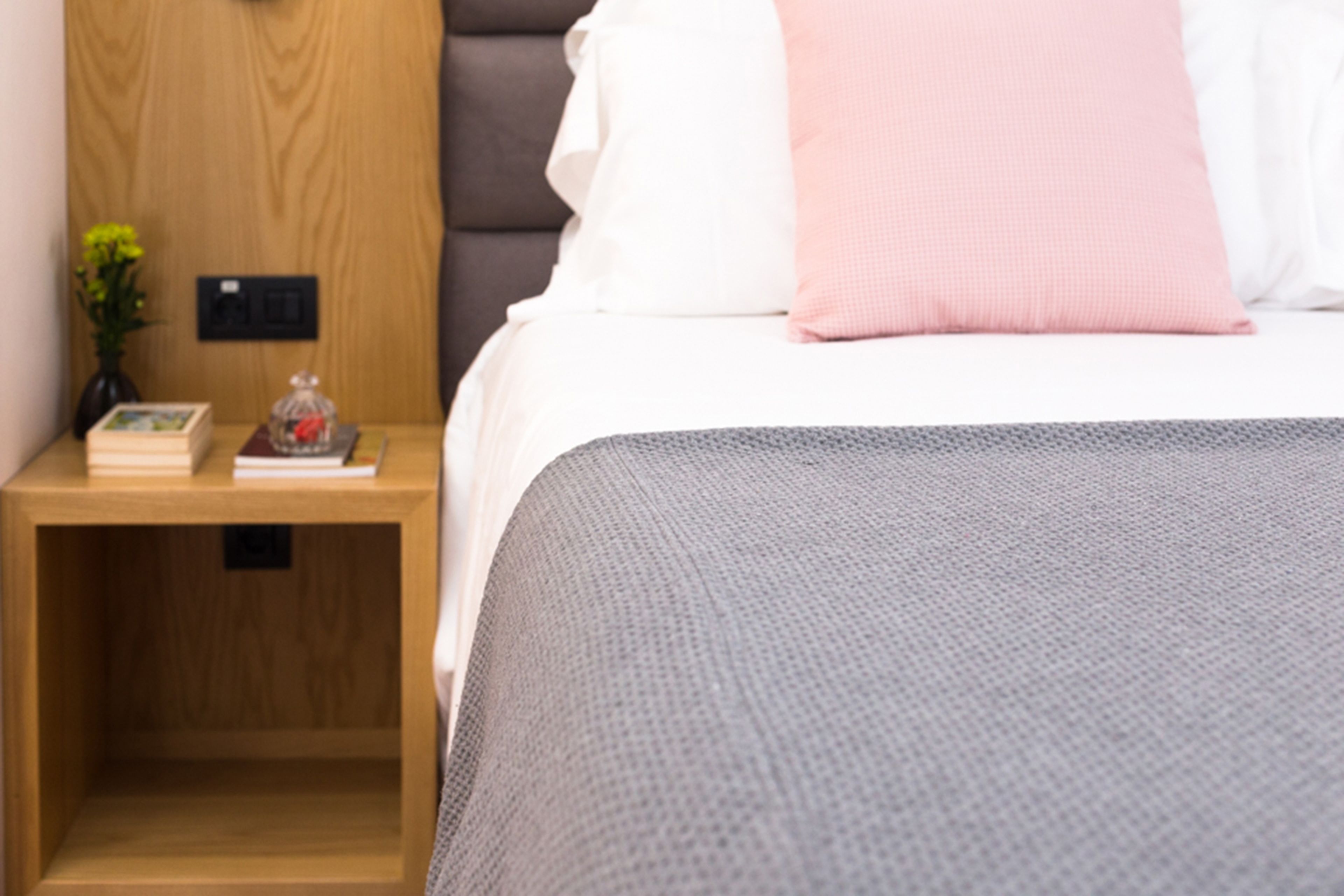 A neatly made bed with a grey blanket, a wooden nightstand with a book and a vase of flowers, and a wooden headboard.