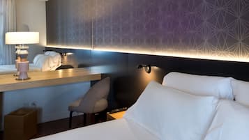 Ett hotellrum med en säng, ett skrivbord med en lampa, en stol och en vägg med geometriska mönster.