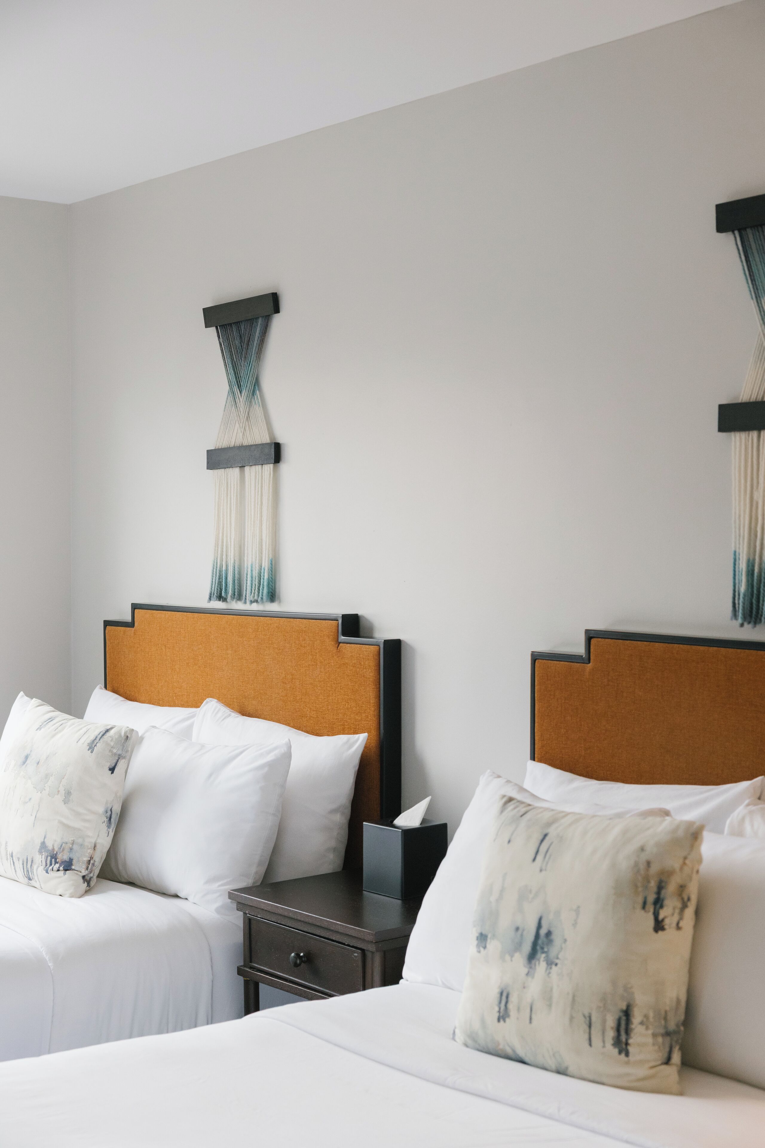A hotel room with two beds, each with white bedding and decorative pillows. There is a small bedside table with a tissue box and a lamp.